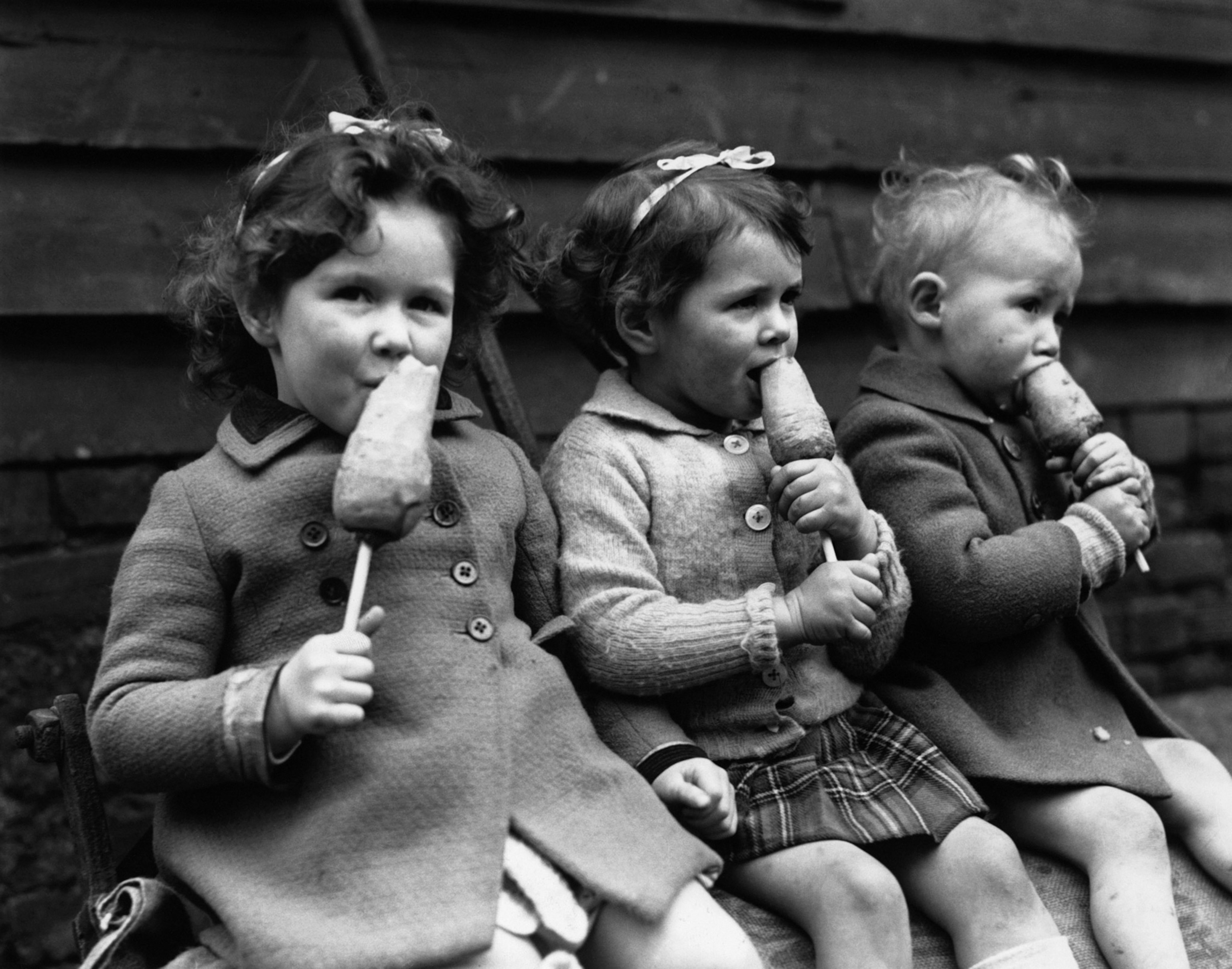 Black and white, three young children sit side-by-side eating a snack of carrots on wooden sticks.
