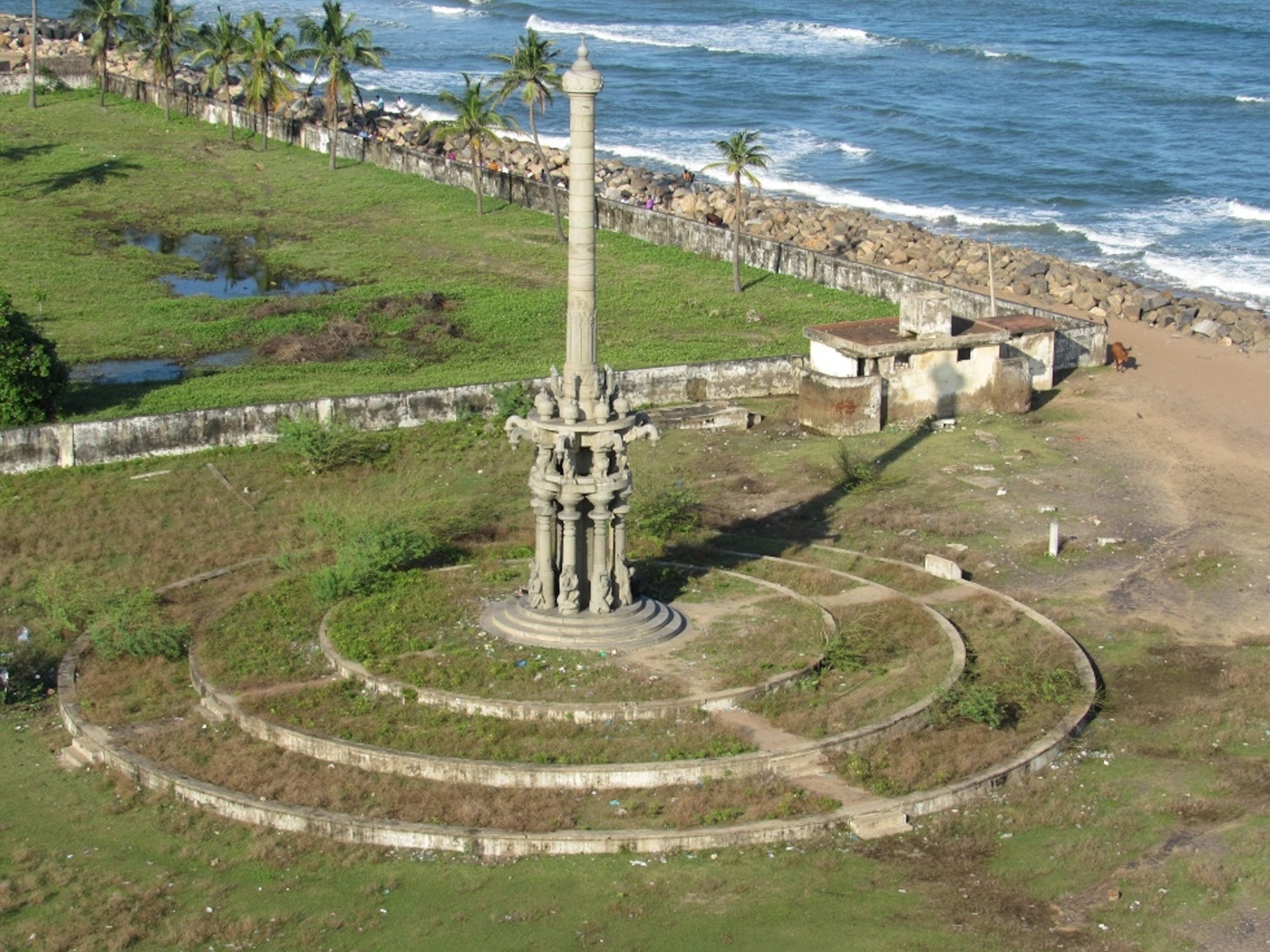 A tall stone monument stands on a circular platform near the shoreline, with palm trees, grassy fields, and ocean waves stretching along the coast behind it.