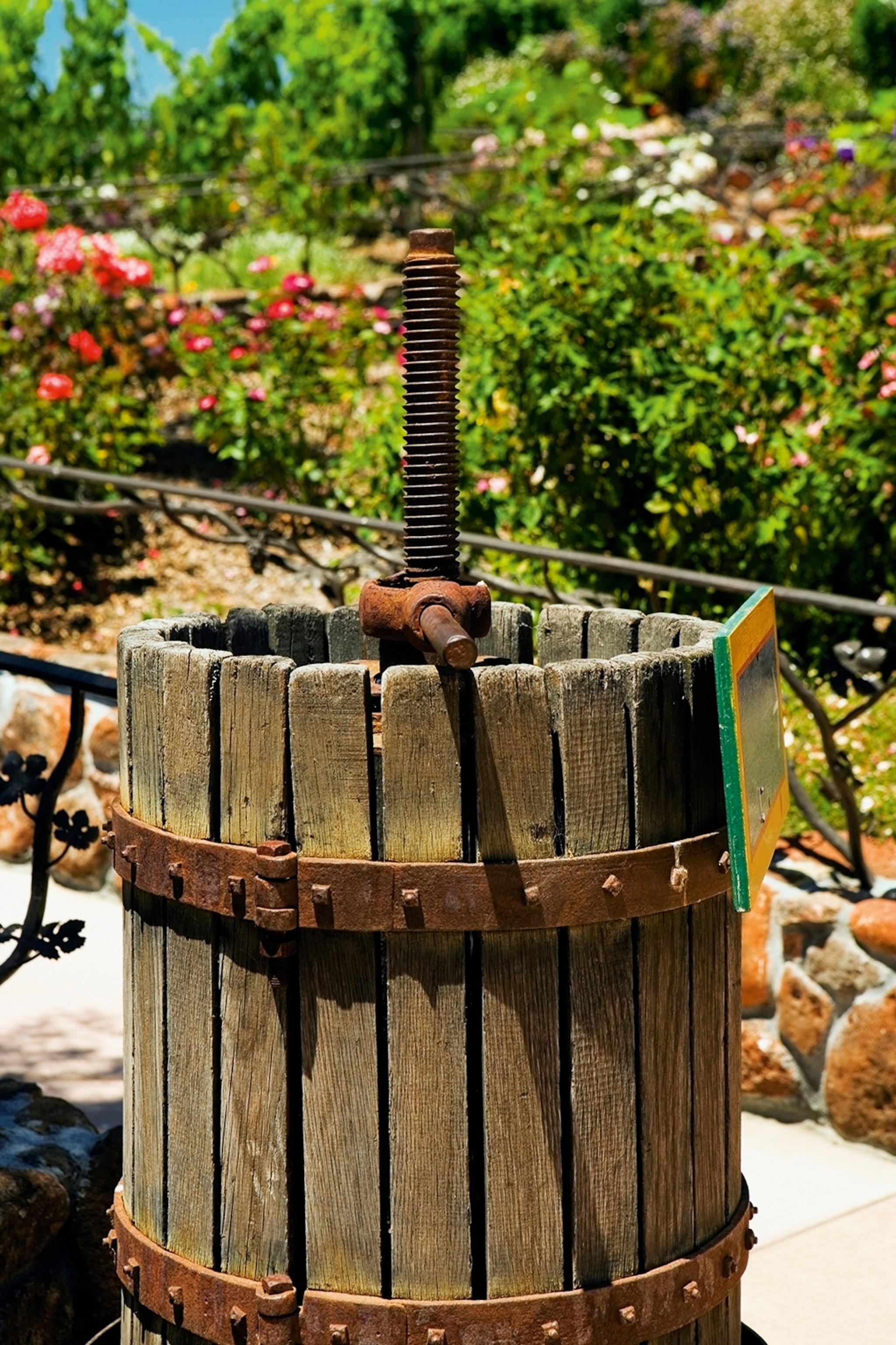 A traditional grape press in the shape of a wooden barrel with a metal press in a lush garden.