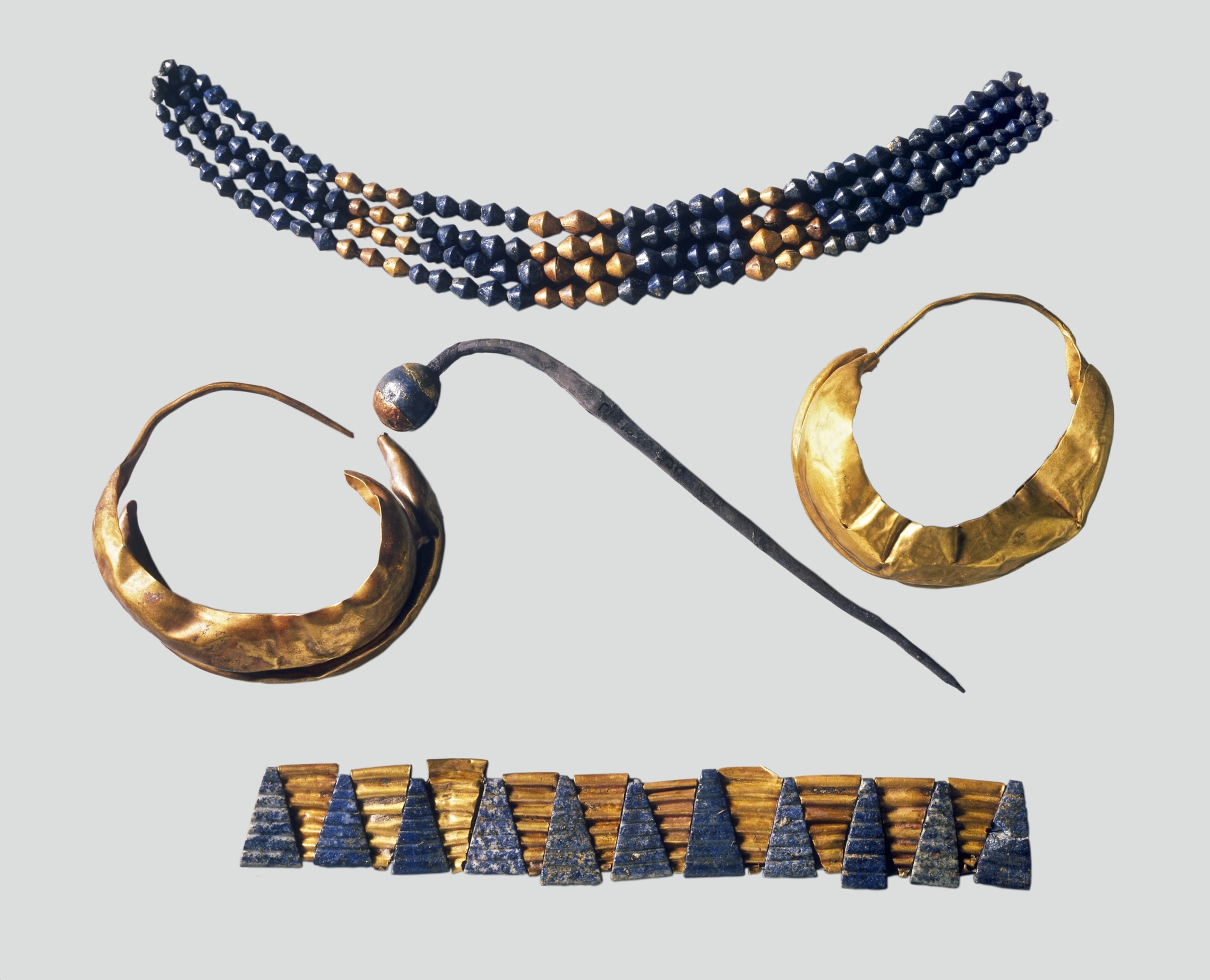 Ancient jewelry set featuring a blue and gold beaded necklace, two gold hoop earrings, a gold and blue patterned belt, and a metal hook, against a plain background