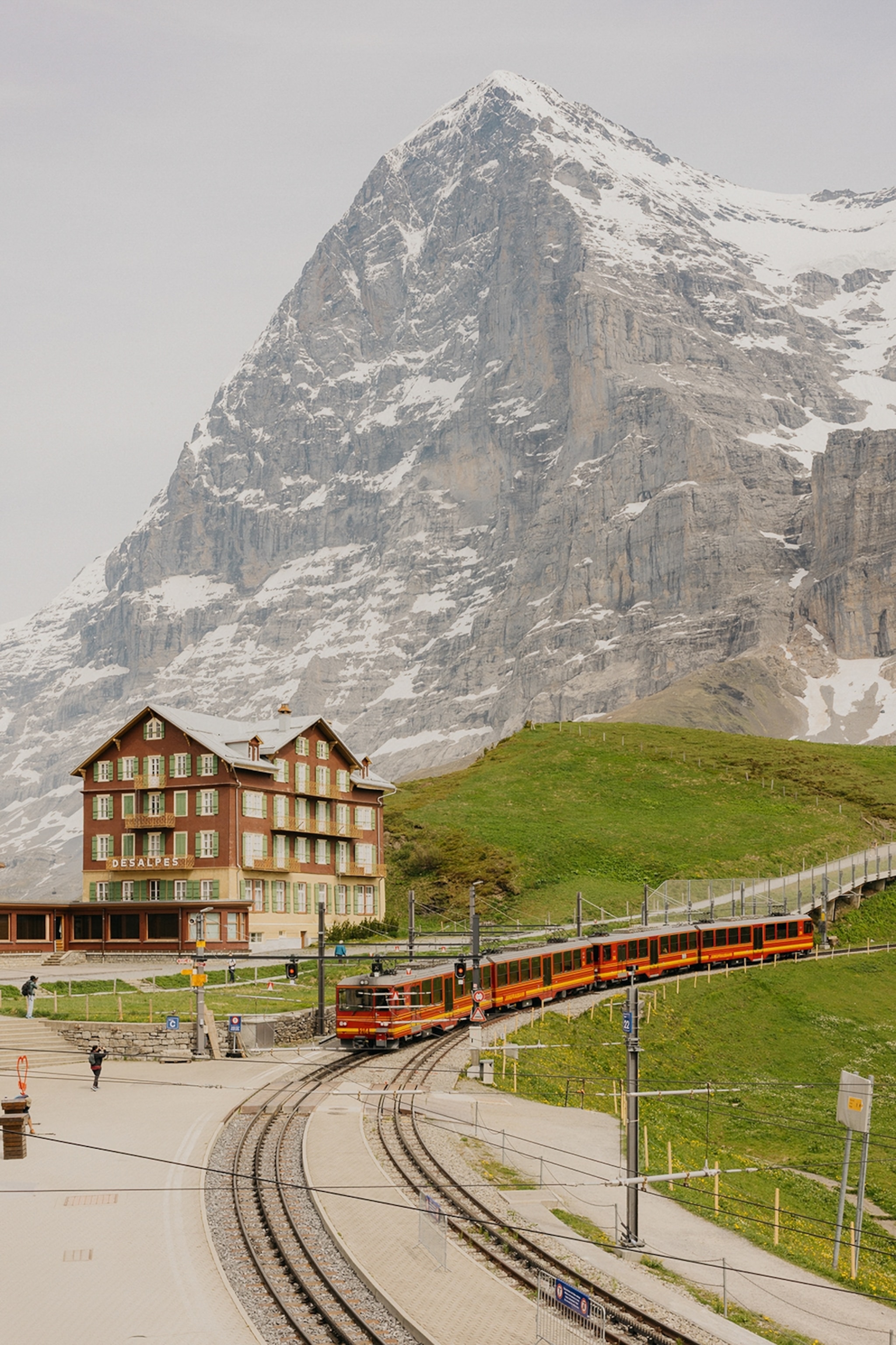 A mountain train coming down a gentle hill with impressive mountains in the background and a traditional house to the side.