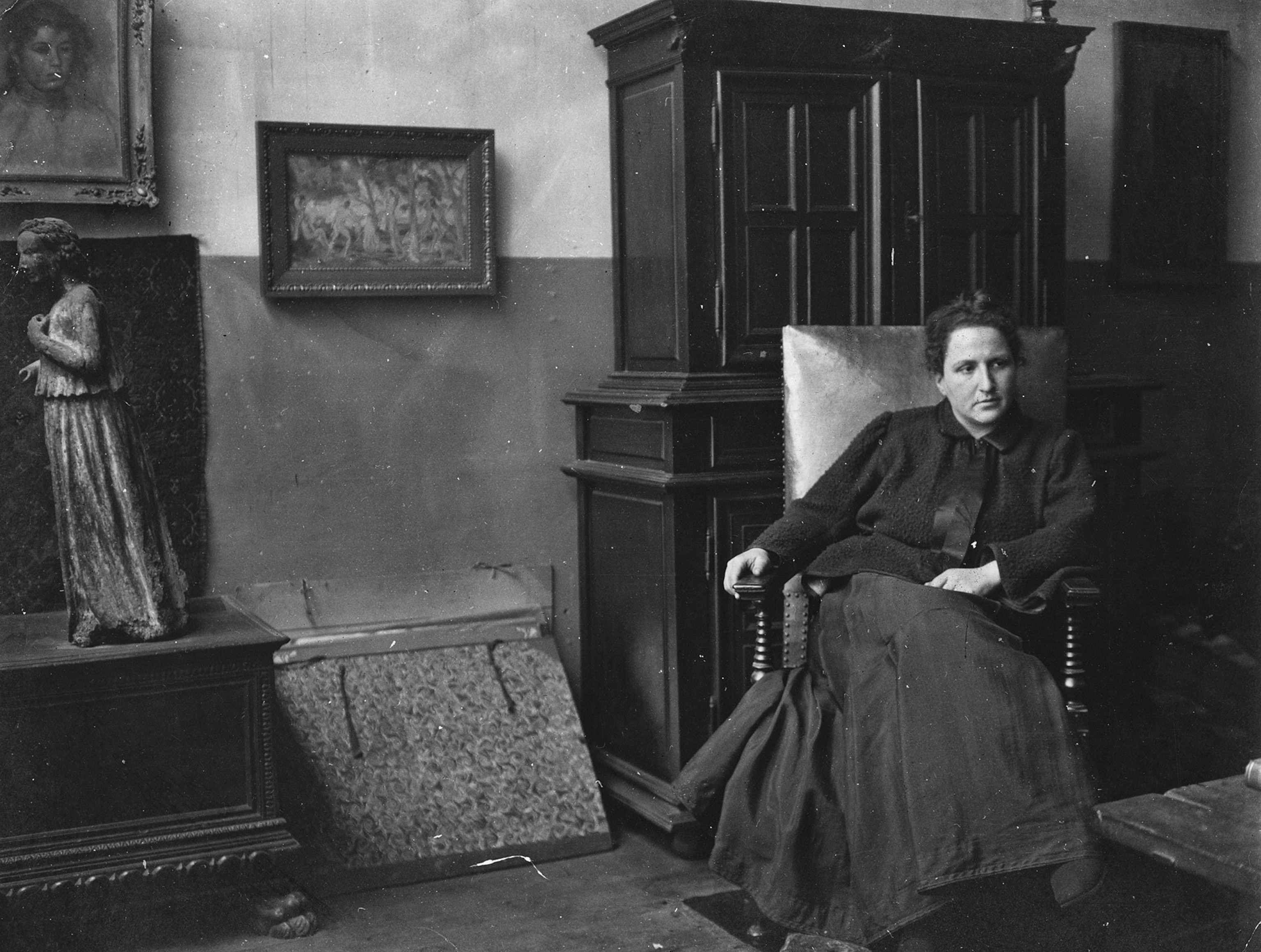 A black and white photo of a woman sitting in a chair in a living area from the early 1900s
