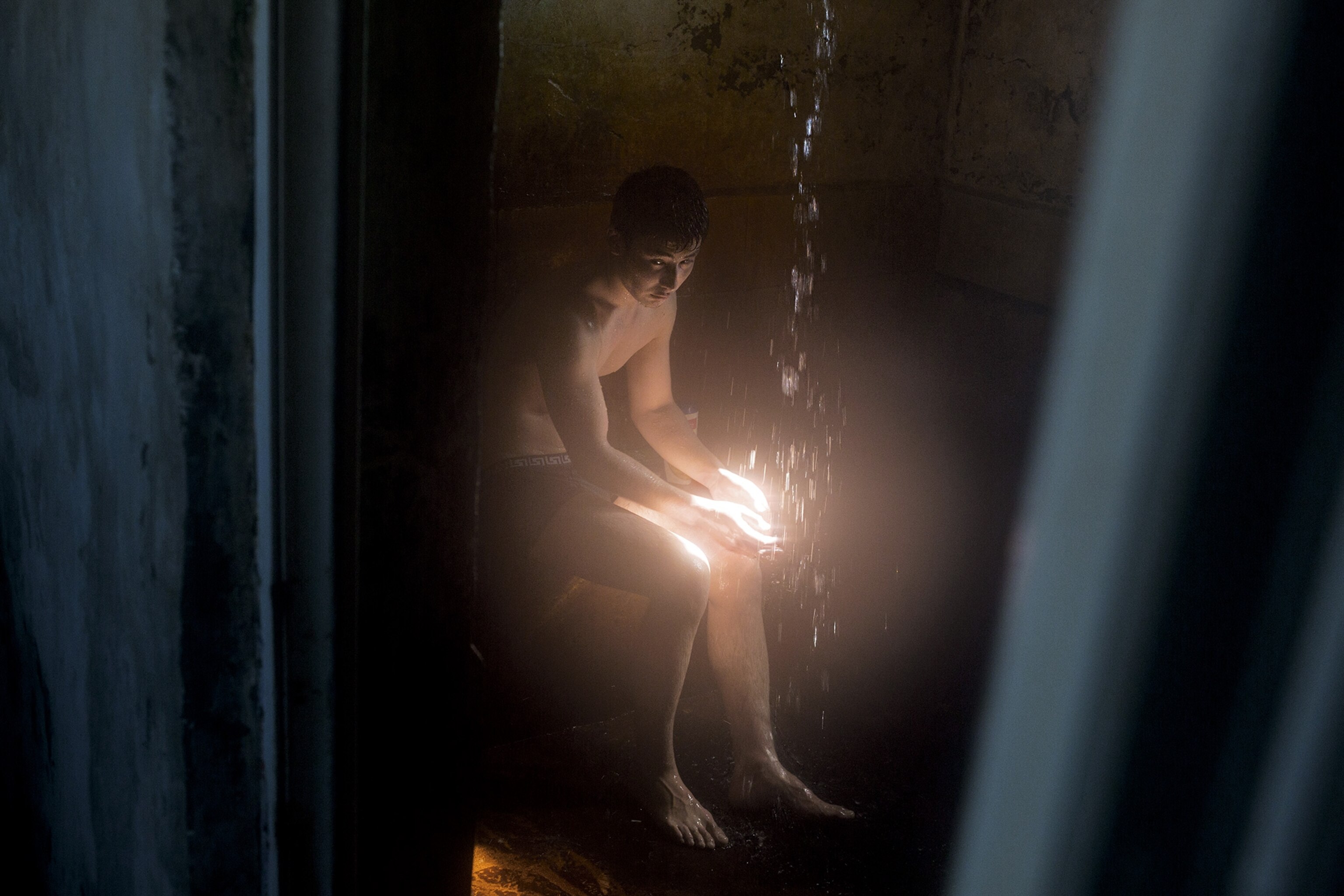 A villager sits inside a bathhouse in Kazakhstan, a beam of light shining on his hands