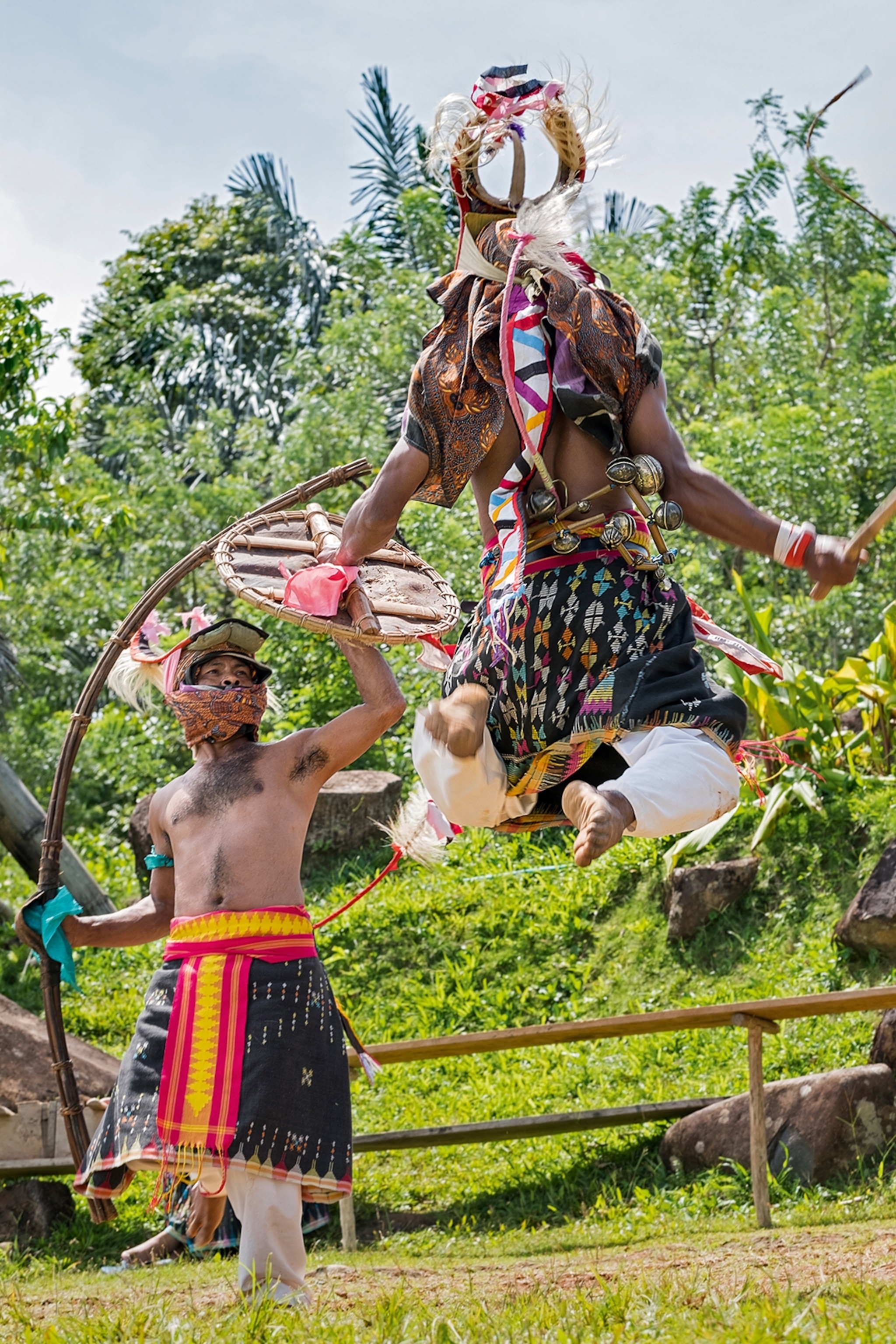 Two local men dressed in hip-high skirts and protective headwear performing a dance with wooden whips, as one jumps into the air.