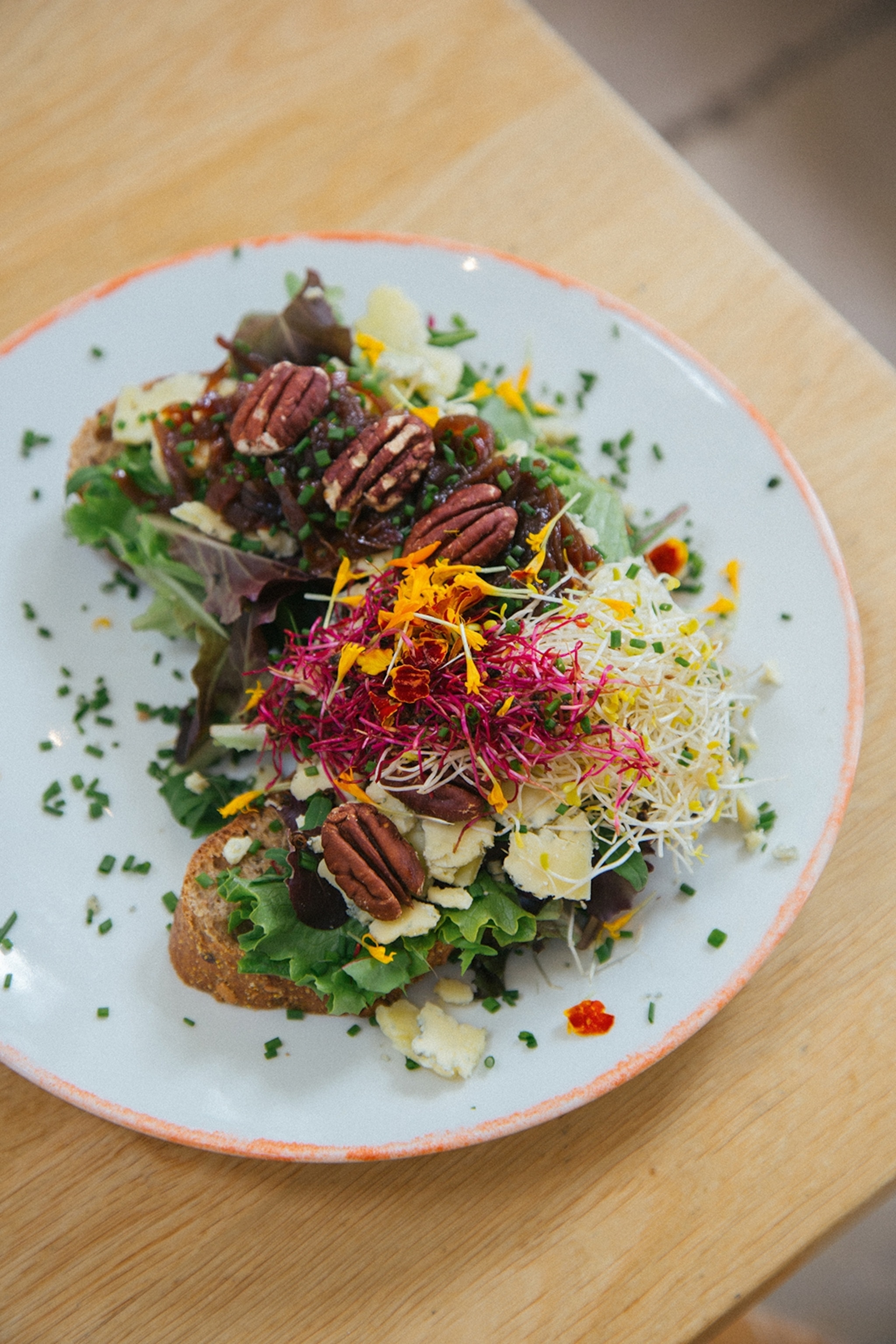 A plate of two slices of toast topped with leaves, cheese shavings, various types of cress, chives and pecan nuts.