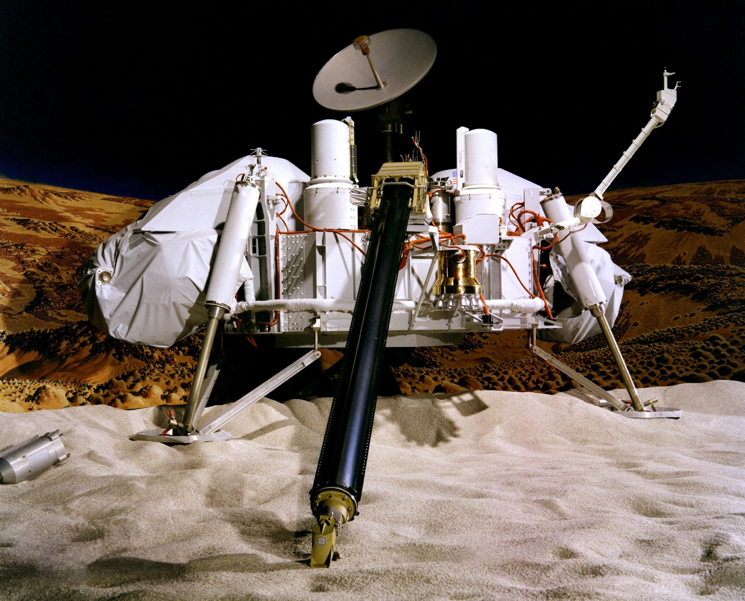 A realistic model of a Mars lander sits on a sandy landscape. It features a large dish antenna, multiple instruments, and mechanical arms.