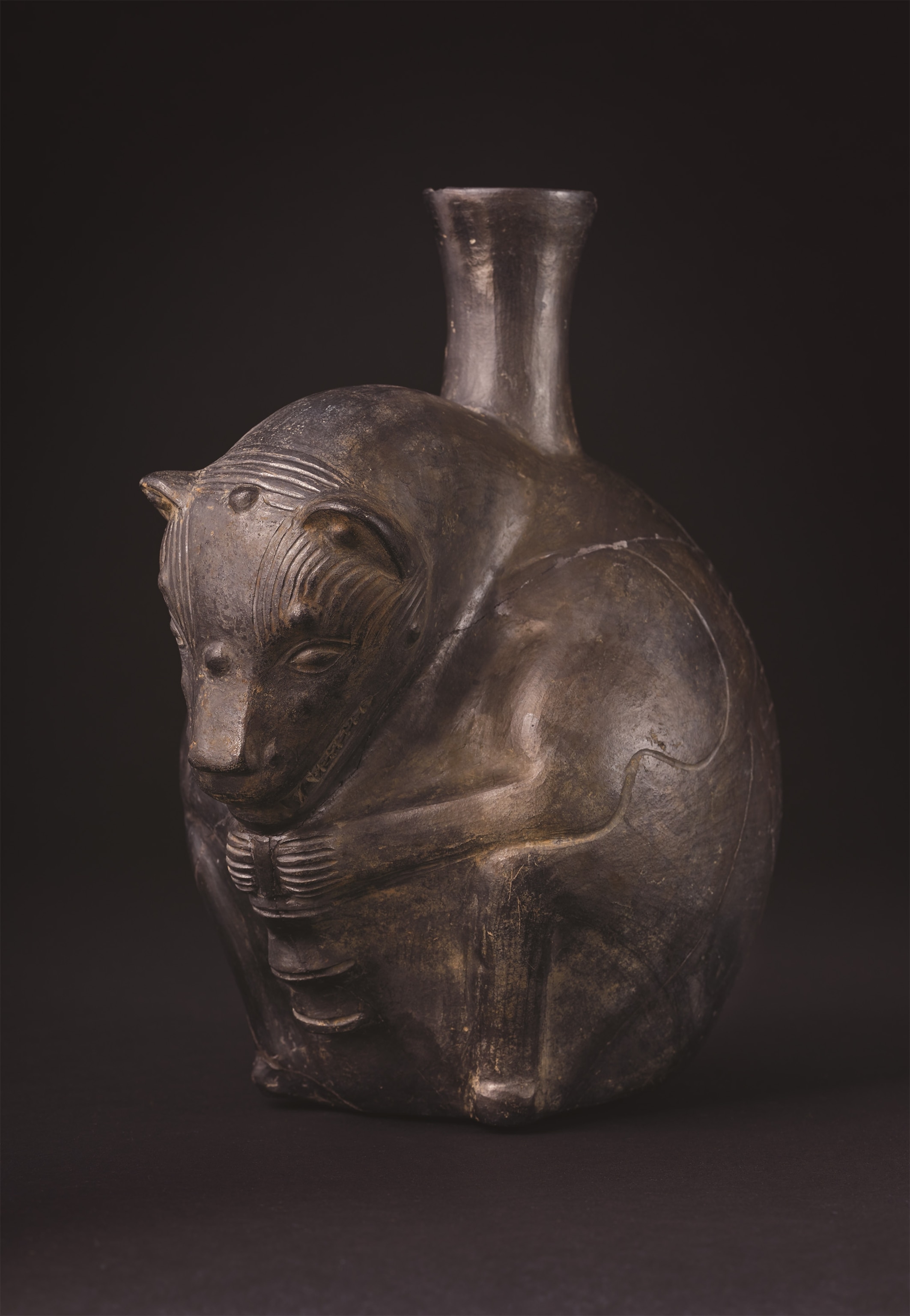Brown and gray earthenware pot depicting a Peruvian hairless dog in a crouched position holding an oblong object, with a spout-like opening protruding from it's back.