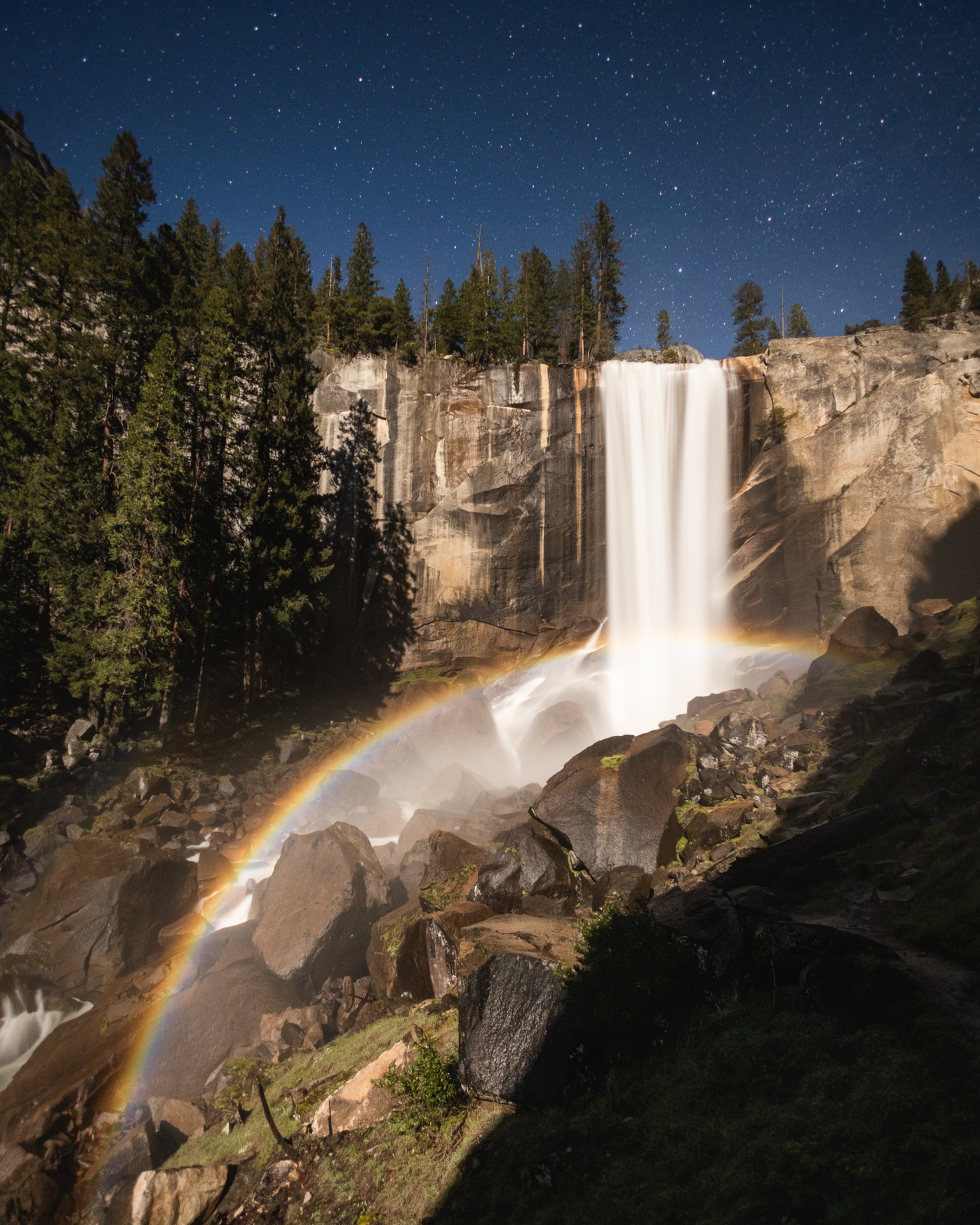 A tall waterfall flows down rocky cliffs at night, with trees above, stars in the sky, and a faint rainbow in the mist near the ground.