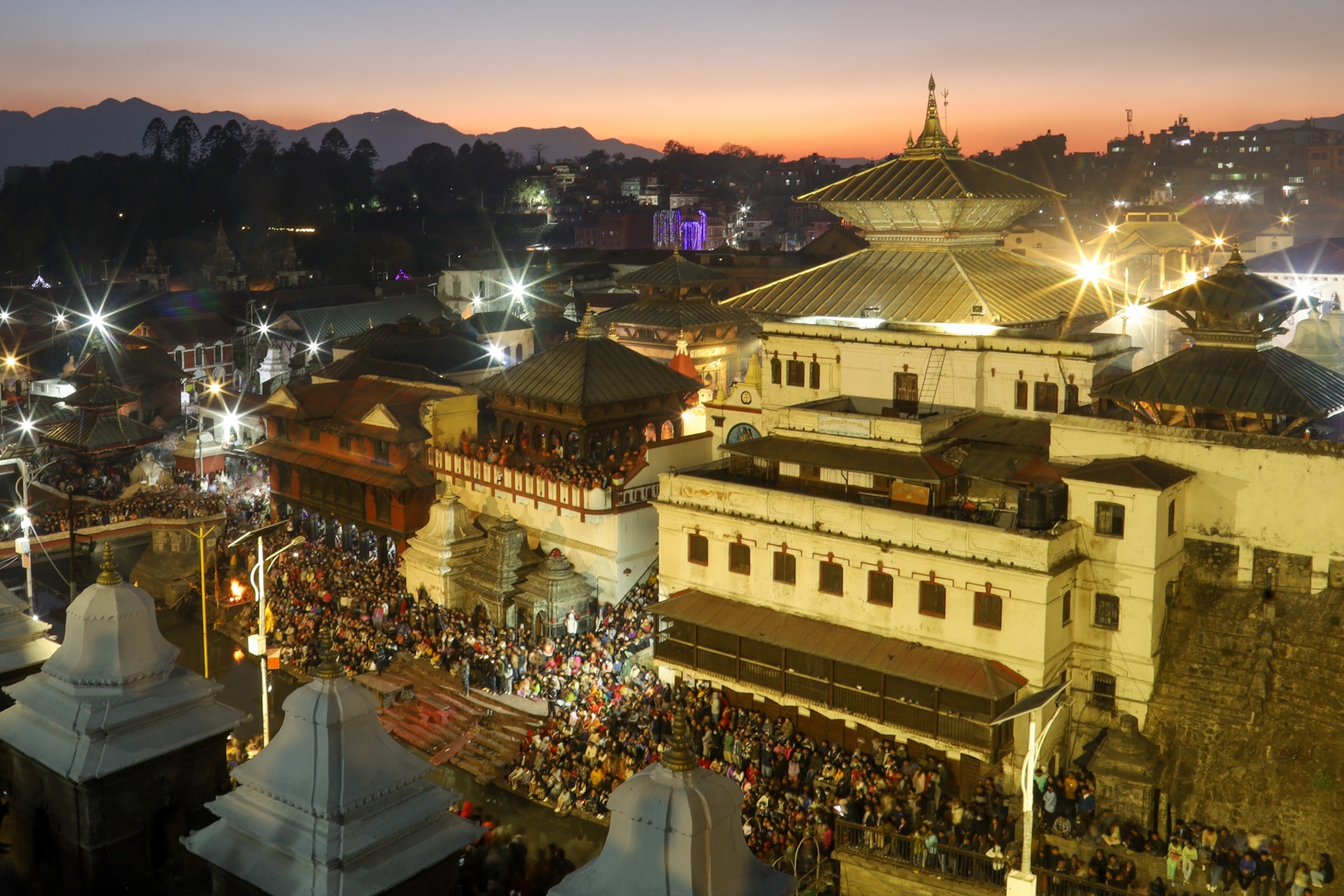 Crowds gather at dusk by Pashupatinath Temple in Kathmandu. Illuminated spires glow under a vibrant sunset, with silhouetted hills in the background.