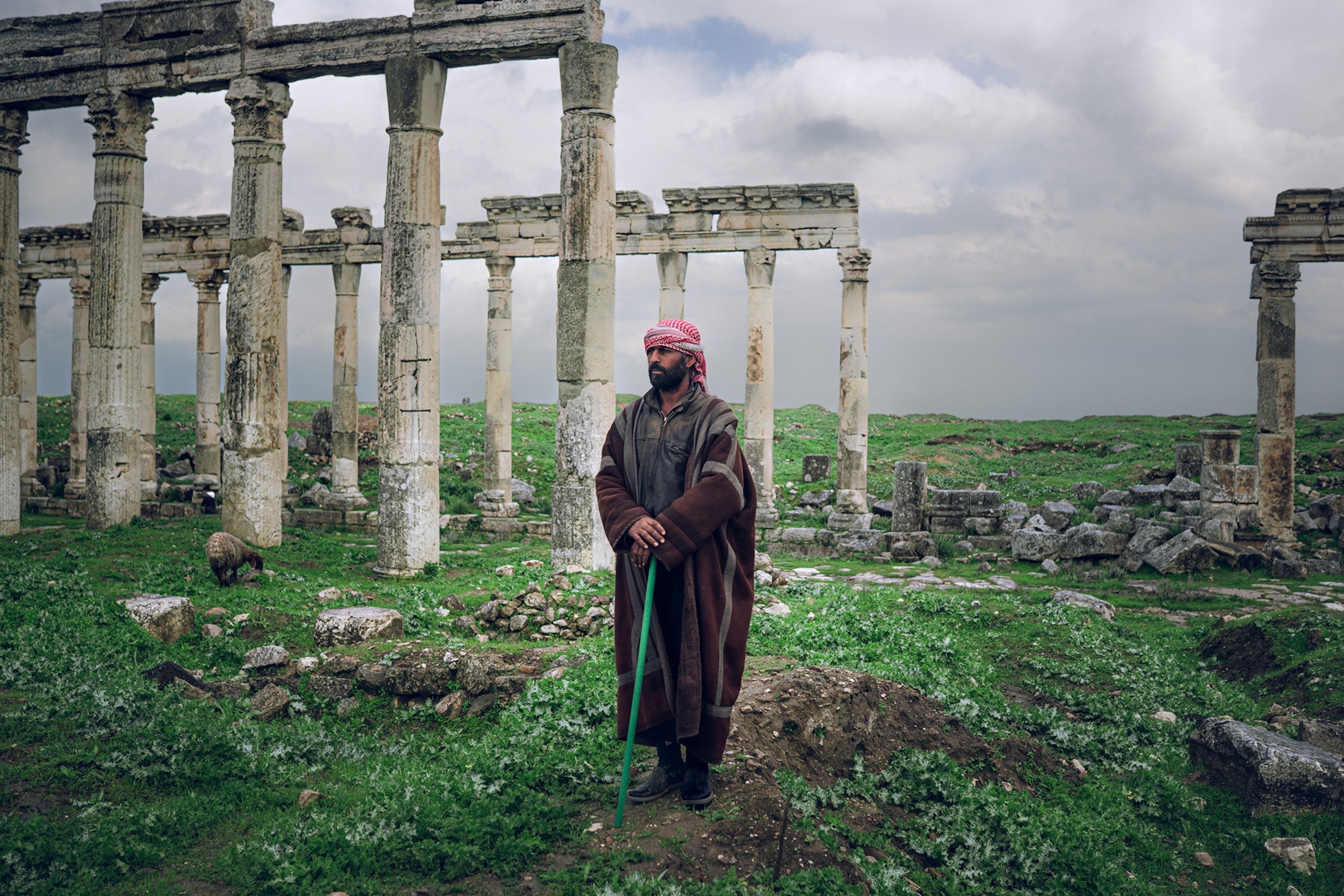 A man with a green cane and a long brown robe stands in front of ancient ruins.