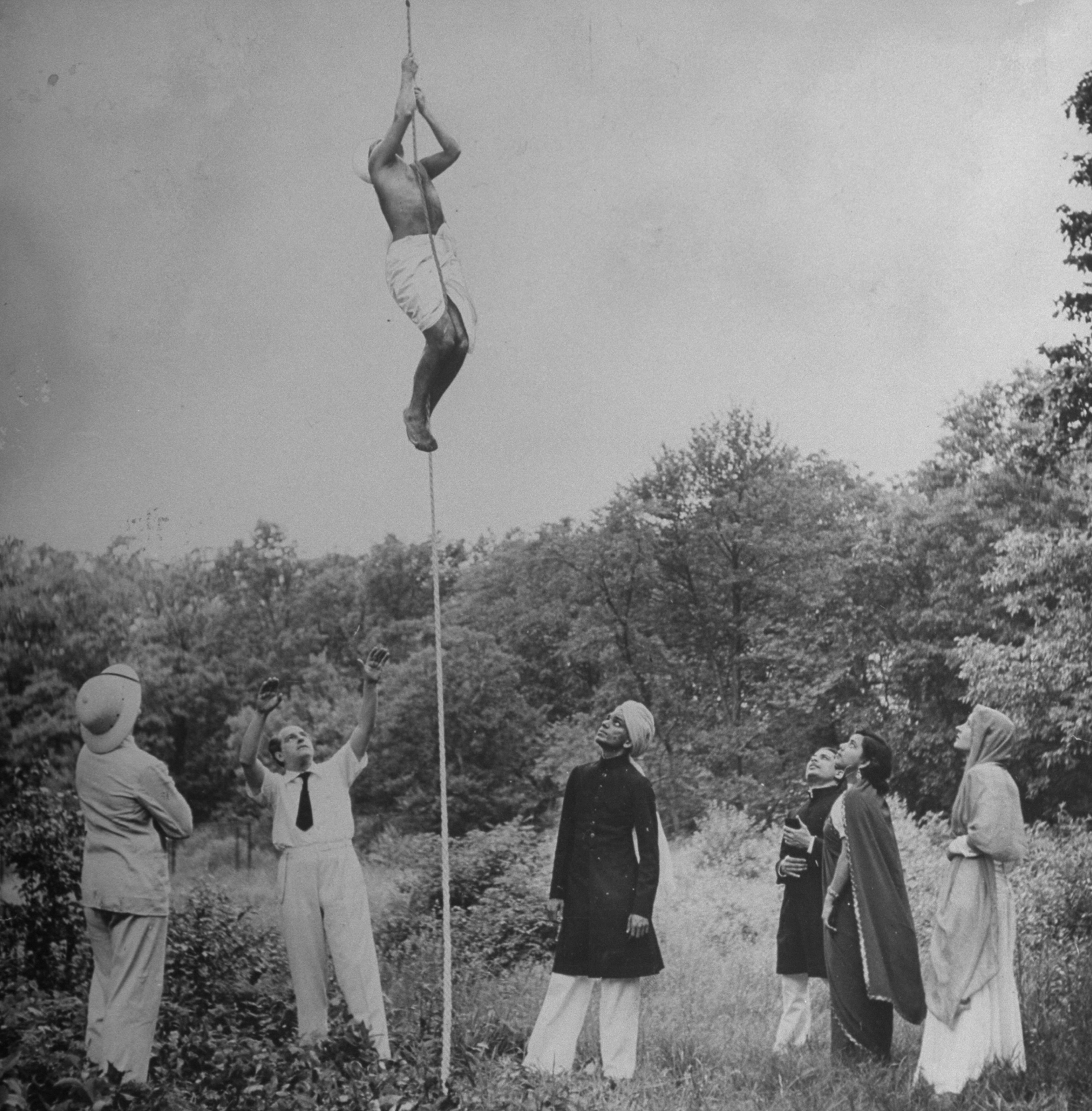 A group with heads upturned as they watch another man who is climbing a rope, which appears to be suspended in mid air