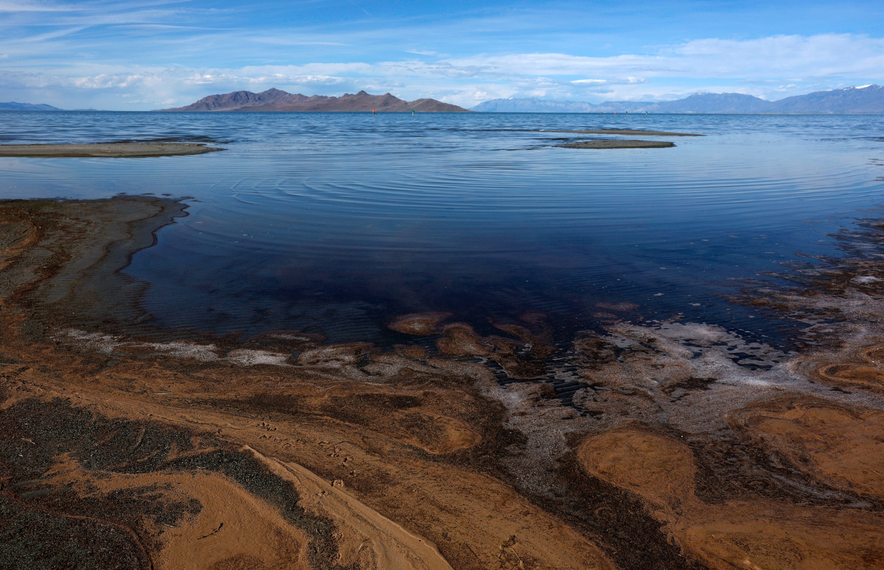 Aerial view of a calm, reflective lake meeting a rocky shoreline with ripples on the water. Mountains and blue sky under scattered clouds in the background