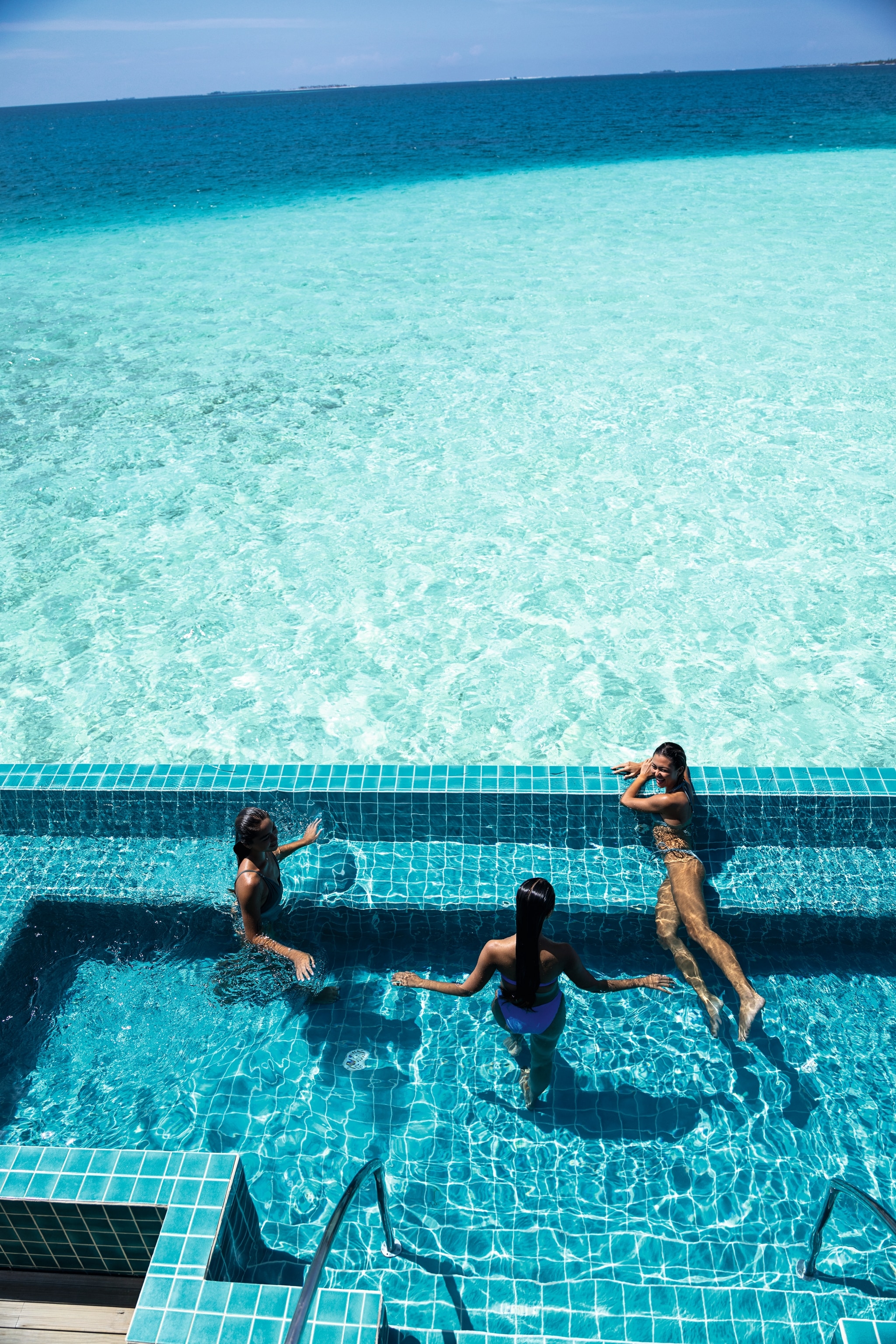 Three people relaxing in blue tiled infinity pool overlooking the ocean. 