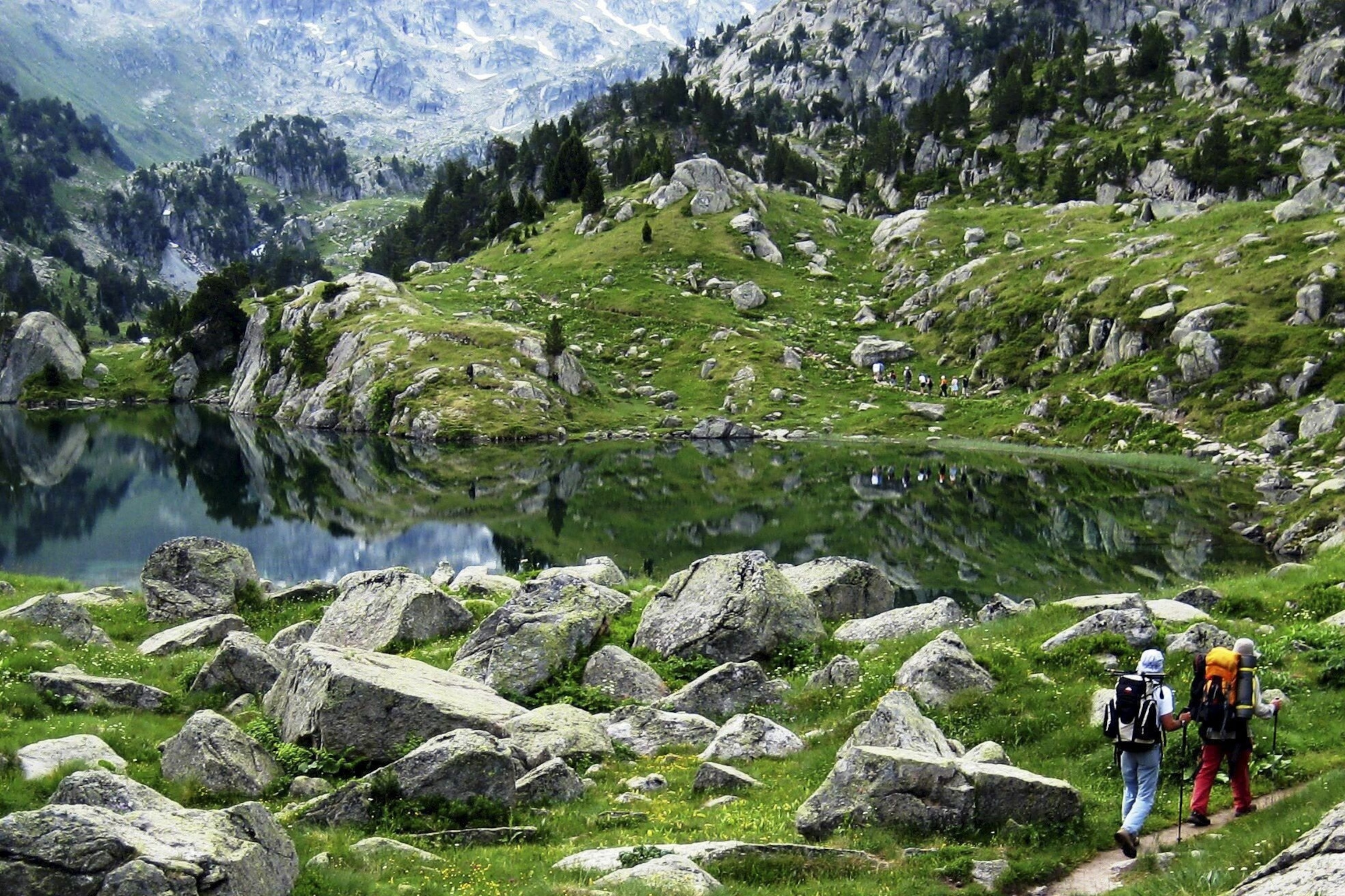 People hike through Aigüestortes i Estany de Sant Maurici National Park, around a glassy river.