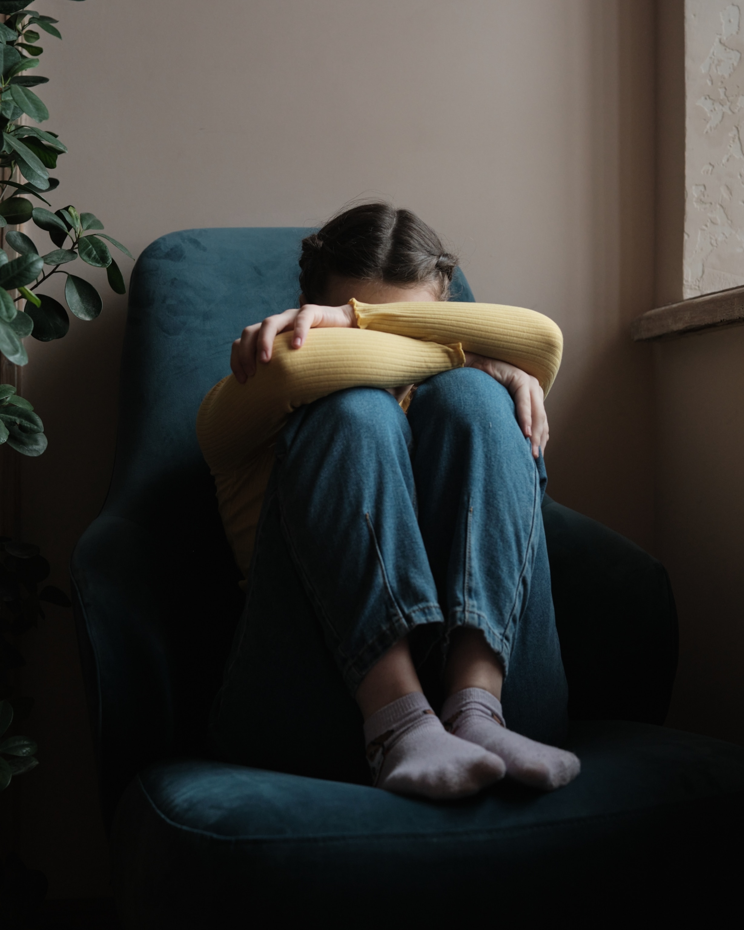 A young teenage girl in a yellow sweater and jeans sitting alone, curled up on a blue armchair, arms wrapped around her knees. The setting is dimly lit, conveying a somber mood.