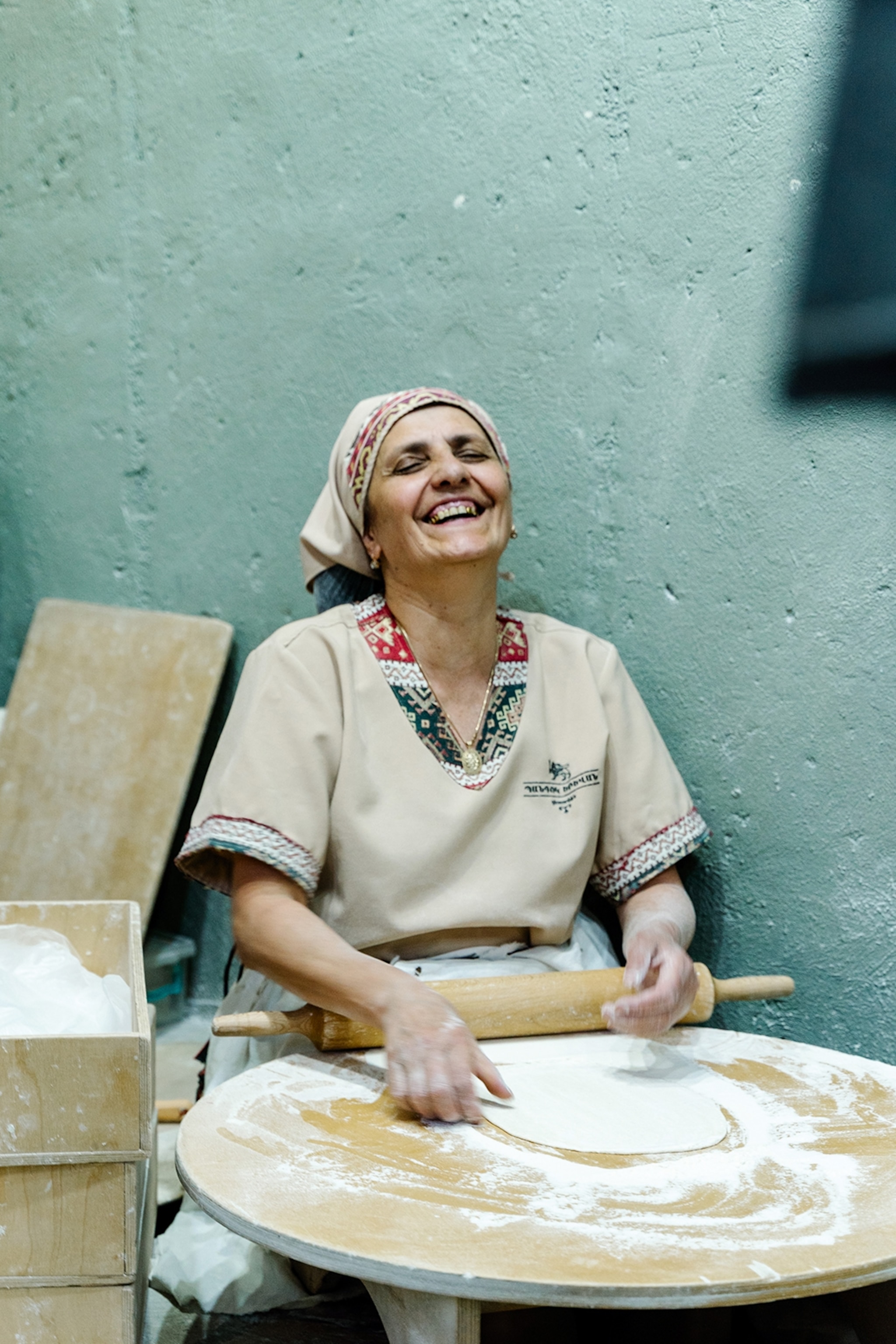 A local woman sat in front of a round, wooden table, shaping a flatbread that's doused in flour, and leaning her head back laughing.