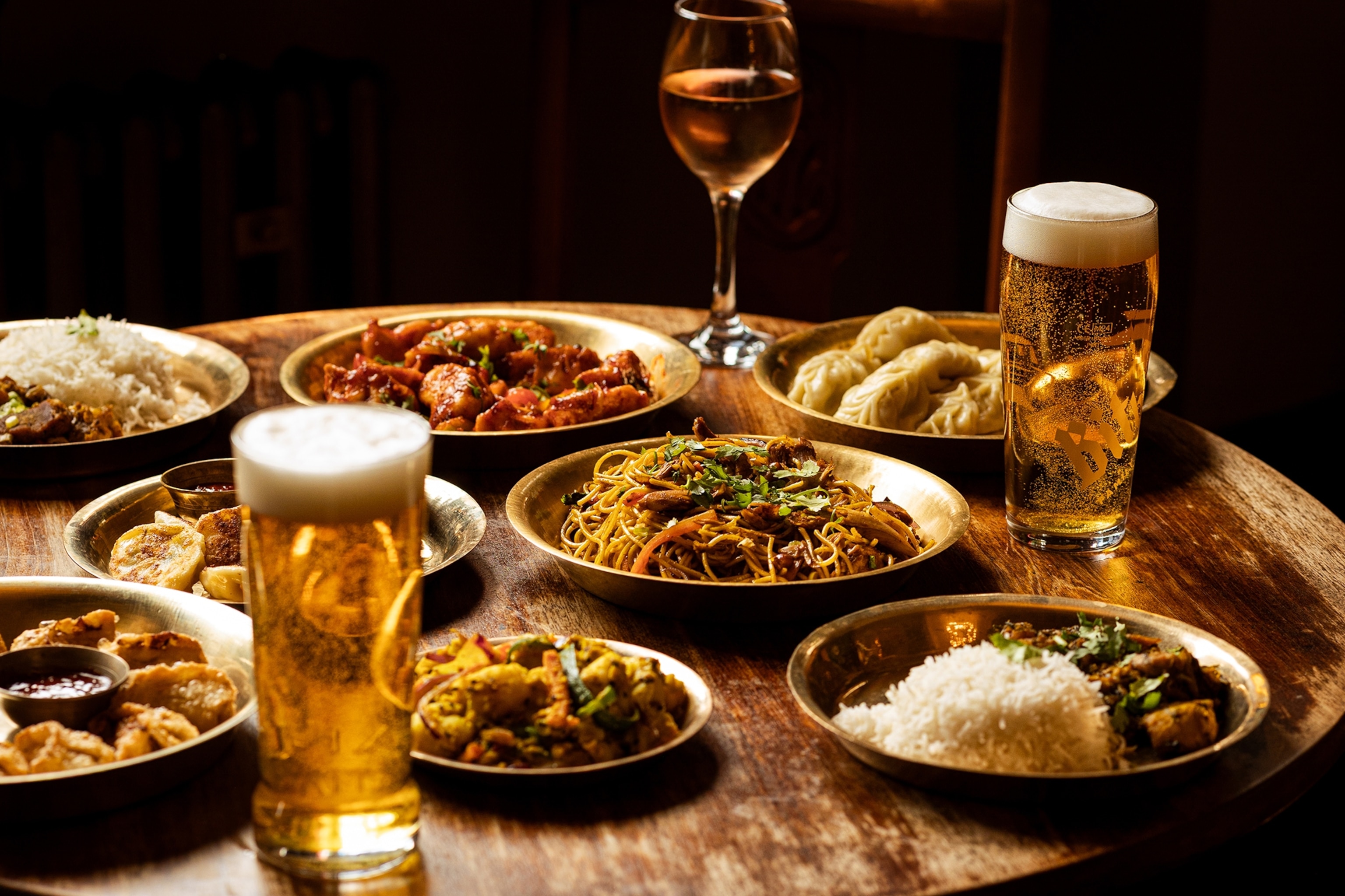 A spread of different small plates holding Nepali street food and dumplings with a glass of wine and pints of beer in between.