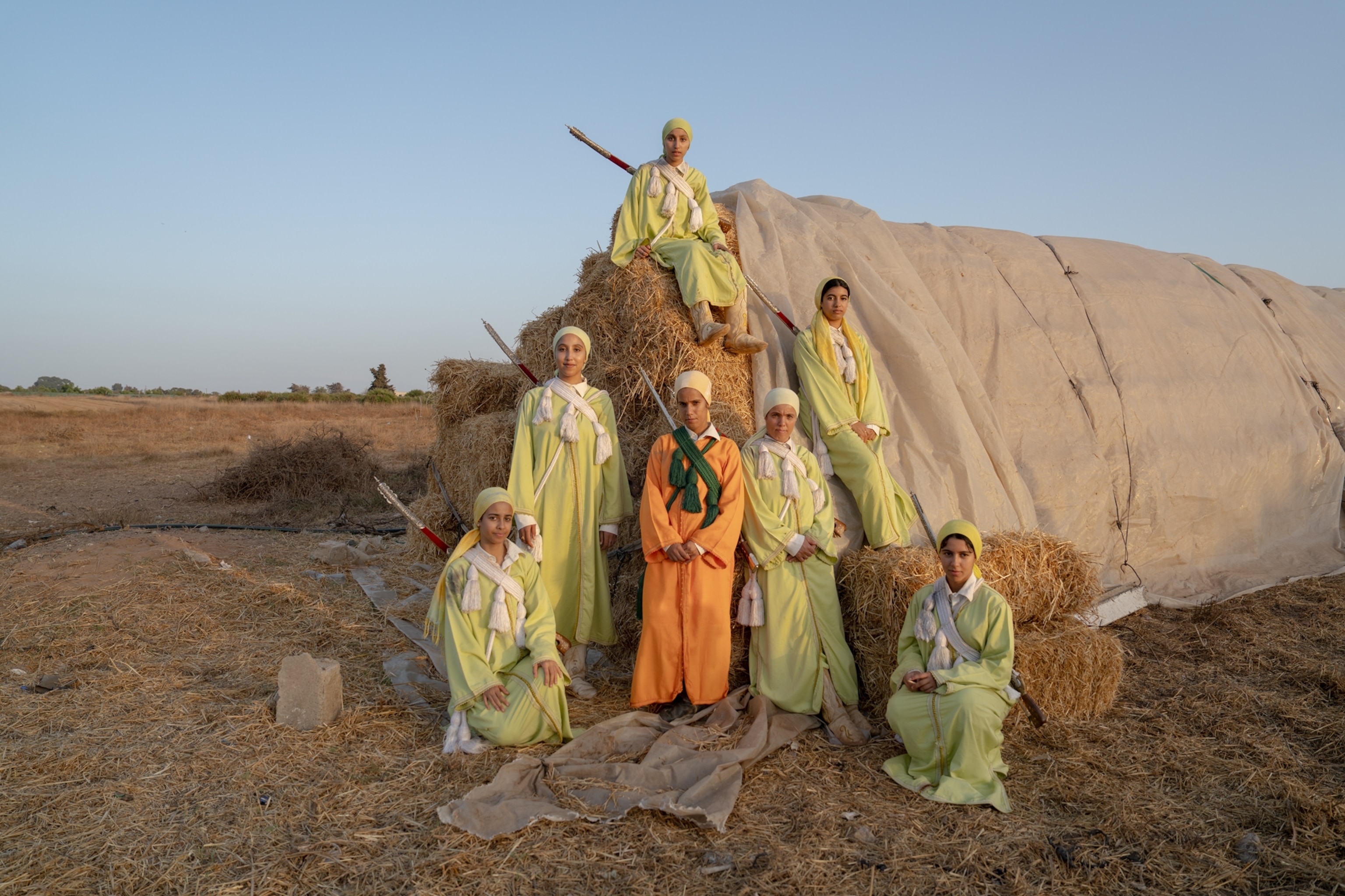 A group of 7 riders sit on hay bails and stand in a pyramid formation. All are wearing bright green, except for the person standing in the middle wearing bright orange.