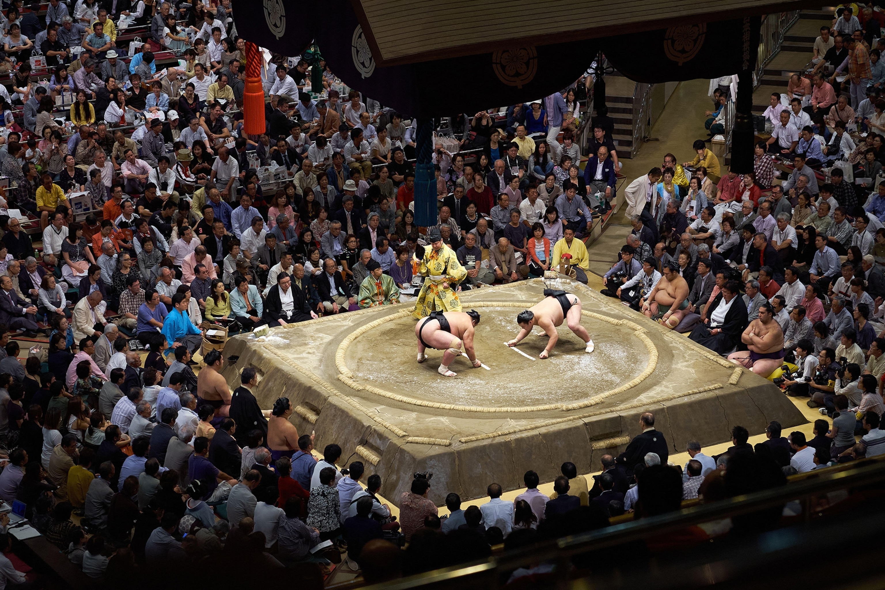 Two sumo wrestlers in the centre of a square platforms surrounded by hundreds of spectators on tribunes.