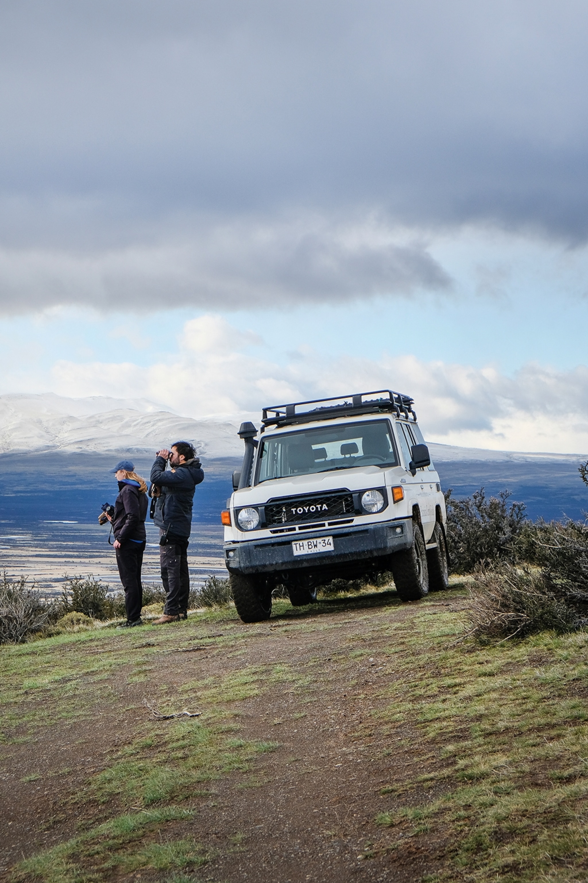 A man and woman in hiking attire standing on the side of a Toyota truck at the top of a mountain.