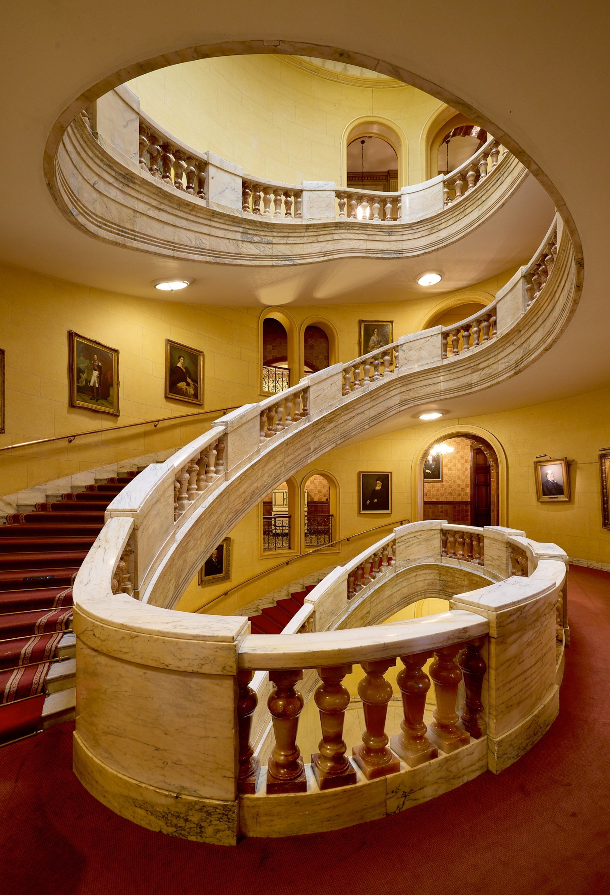 Spiral staircase with ornate marble railing in a grand interior. Warm lighting and portraits on walls create an elegant and historic ambiance.