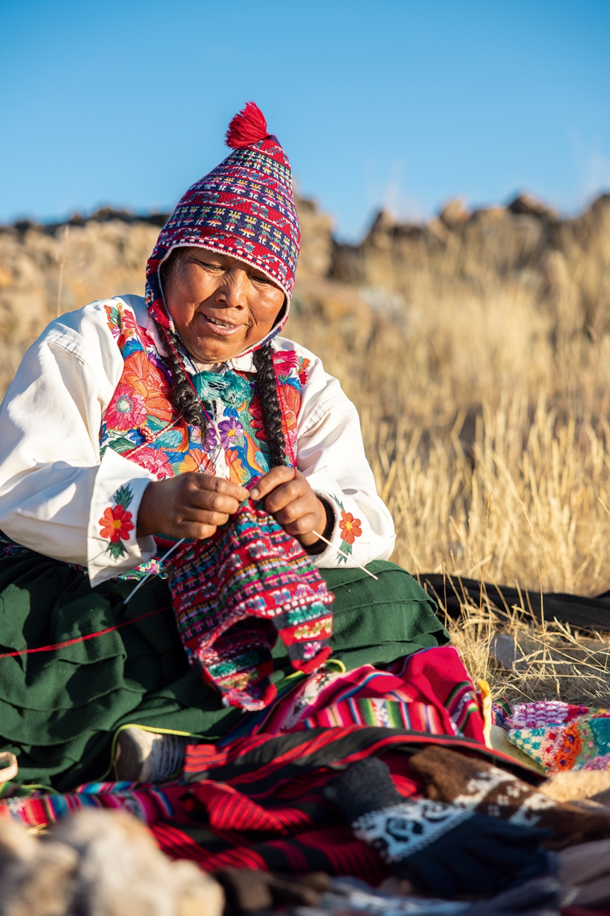 A native Peruvian woman sitting on the ground next to a big lake, weaving a traditional hat.