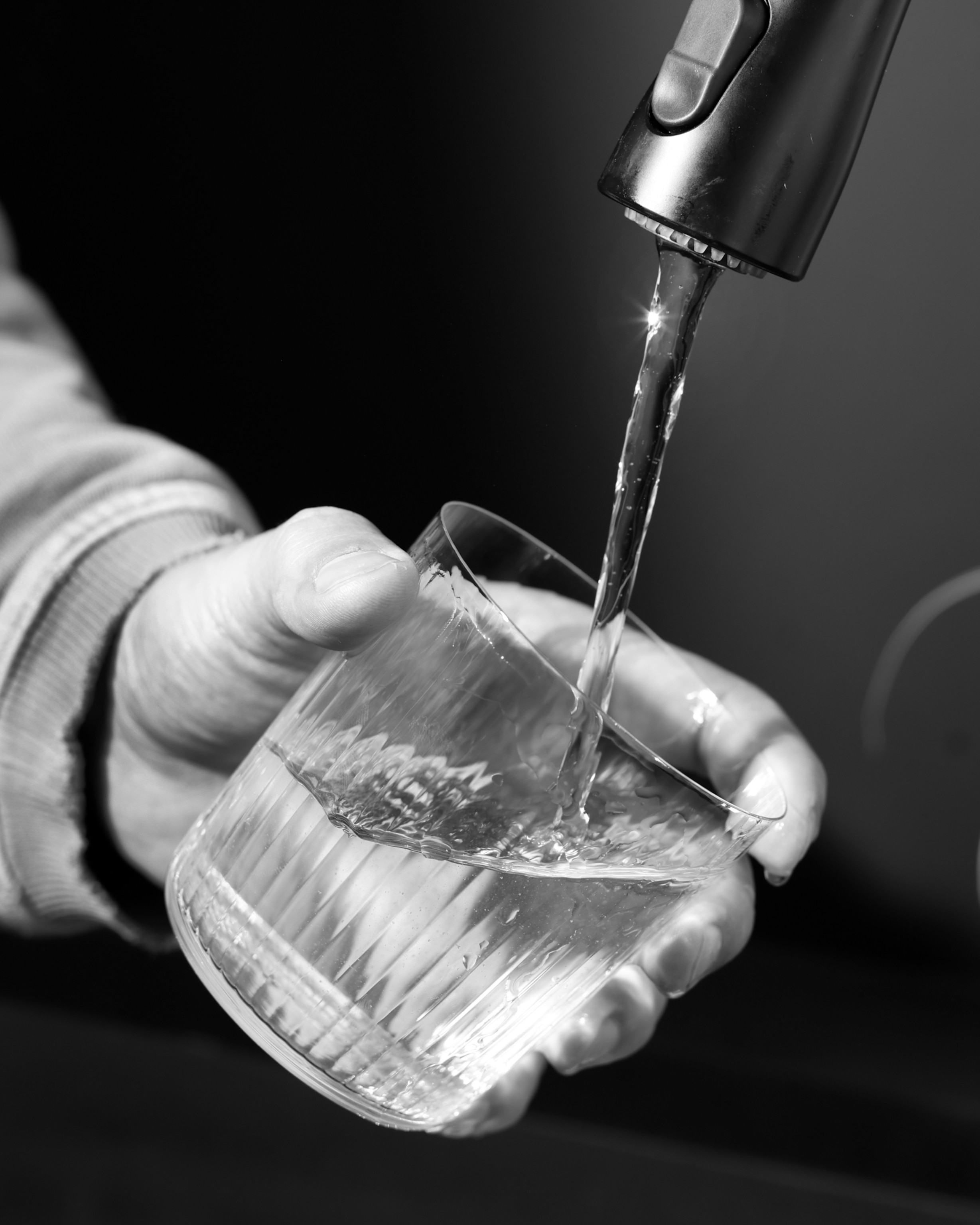 Black and white image of a hand holding a glass under a faucet, with water flowing into it