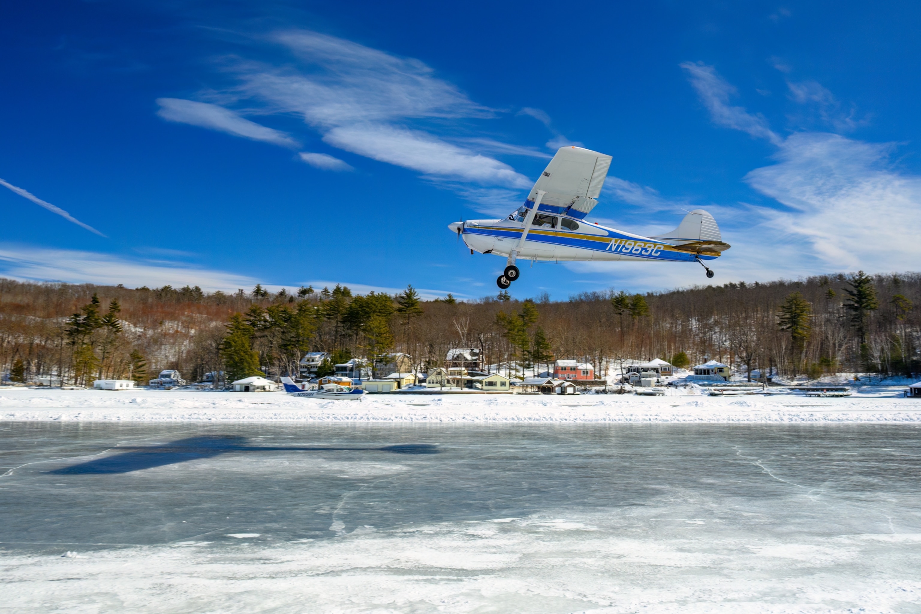 A small plane flies low over a frozen lake, casting a shadow on the icy surface. Snowy trees and houses line the shore under a bright blue sky.