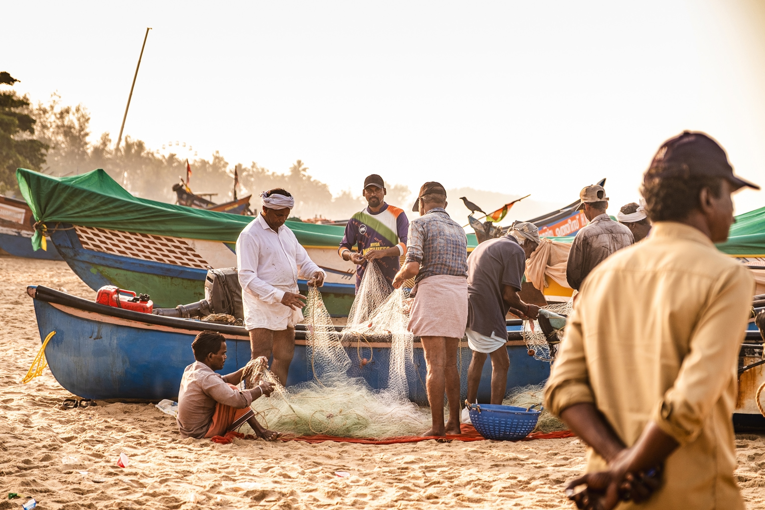 A beach shot at sunrise featuring a group of local fishermen picking small fish from their nets besides lined-up boats.