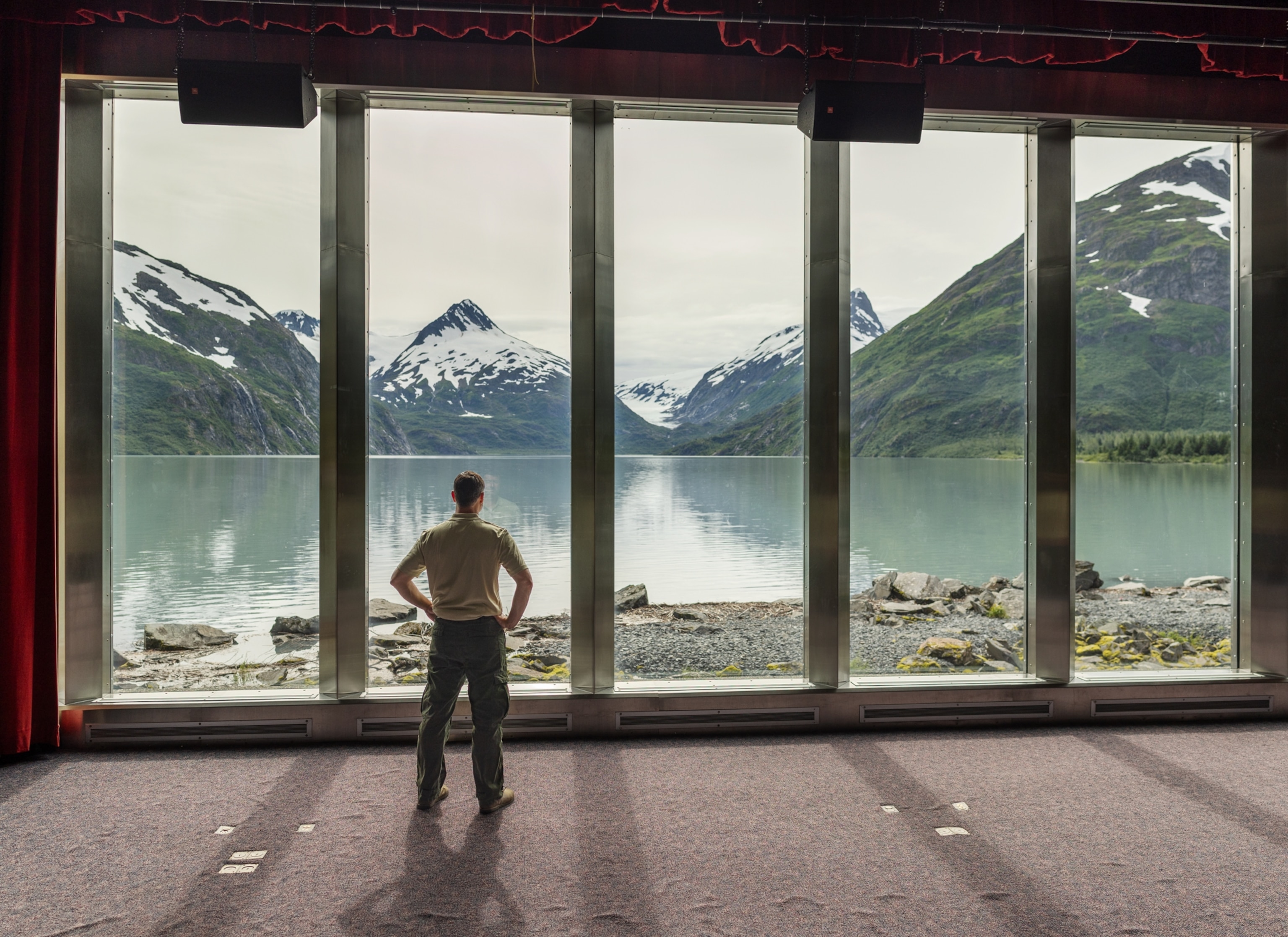 A person stands inside a building, looking out through tall windows at a calm lake and green mountains with snow on their peaks.