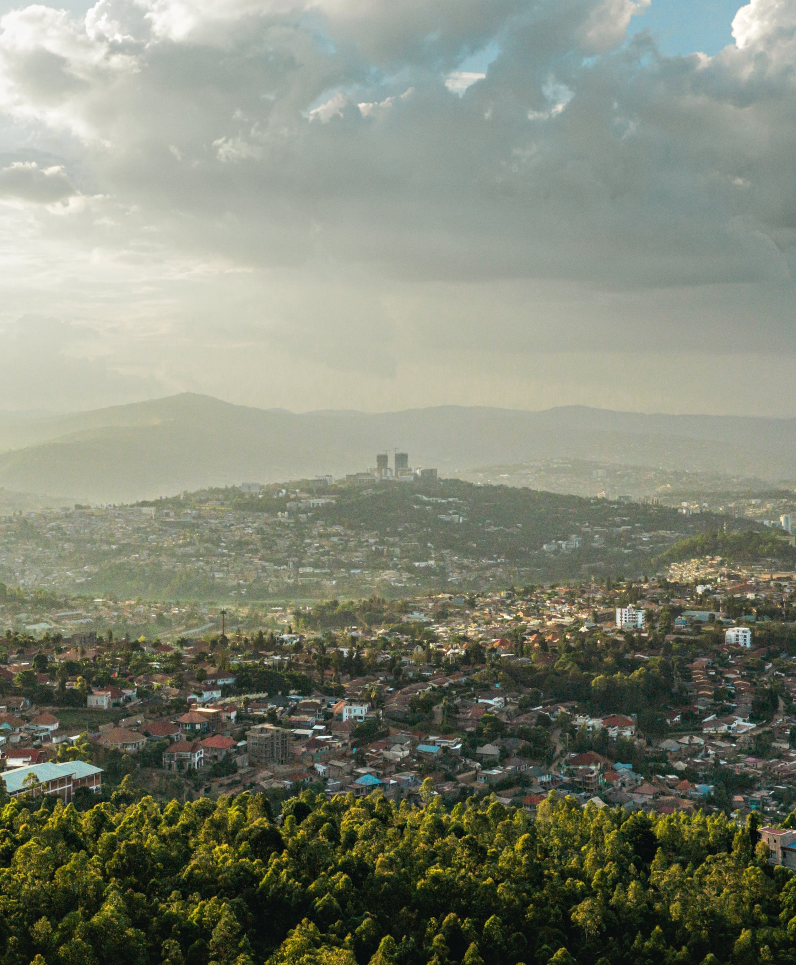 An aerial view of a large city spread across rolling hills and forested areas in the foreground.