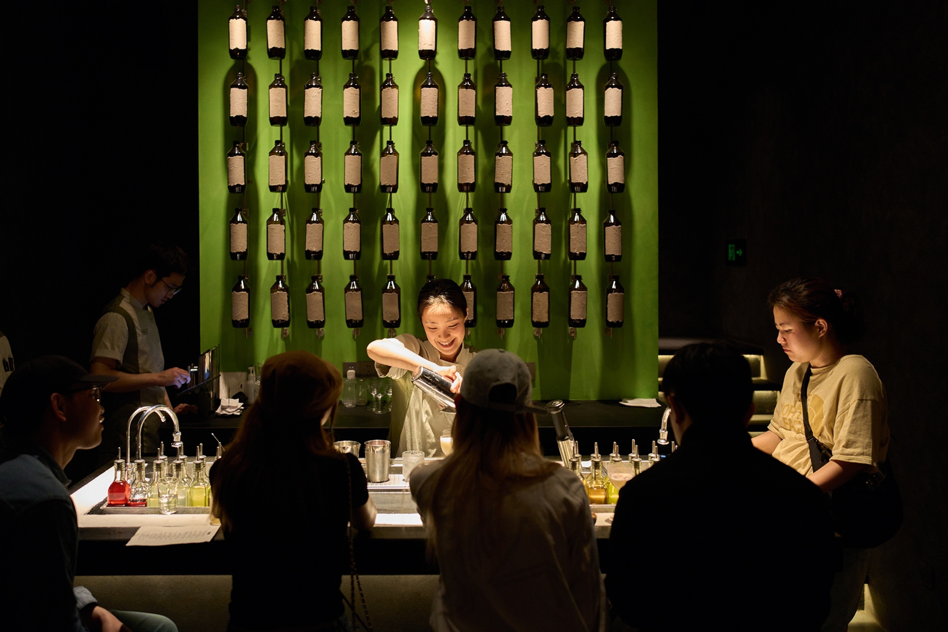 A moody bar with a smiling female bartender pouring a drink to customers in front of a wall with hung bottles.
