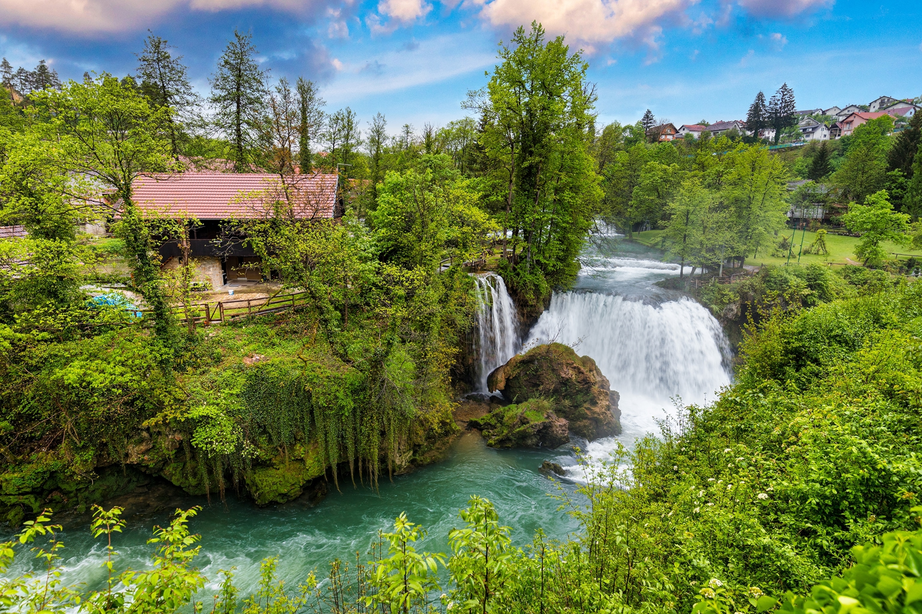 A tall waterfall in the middle of a forest with a village nestled in between trees.