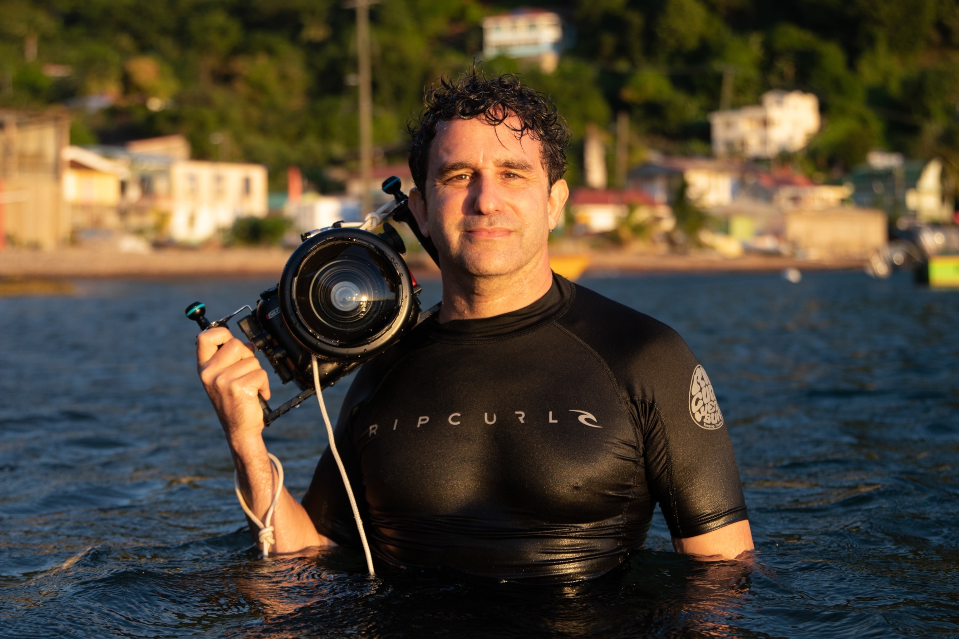 A man with dark hair and light skin, is photographed in a texutred wet suit that fits snuggly, and is holding a large camera with one hand and resting half of the camera body on his shoulder.