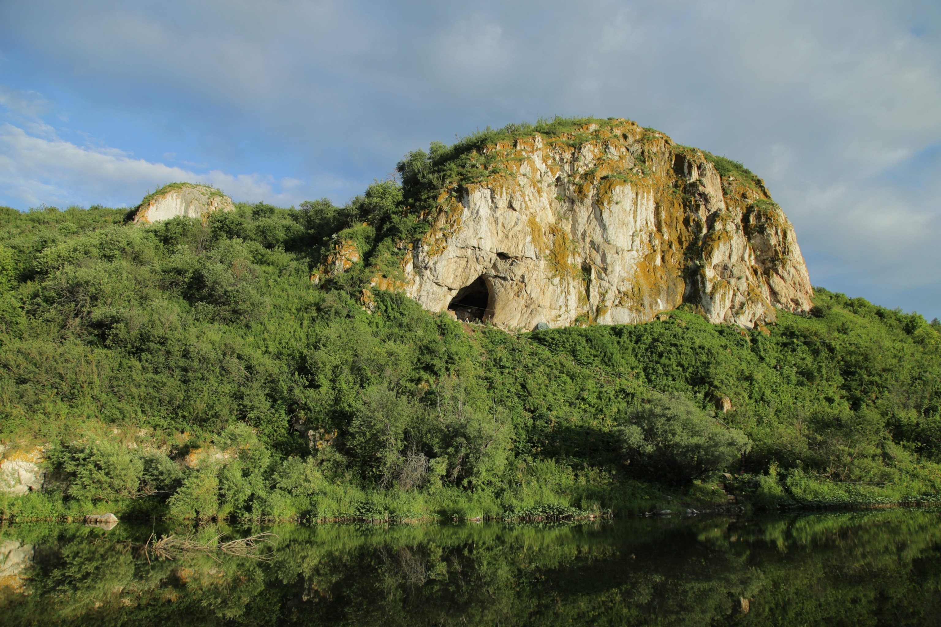 A rocky cliff with a cave entrance above dense green foliage, reflected in a tranquil lake under a partly cloudy sky
