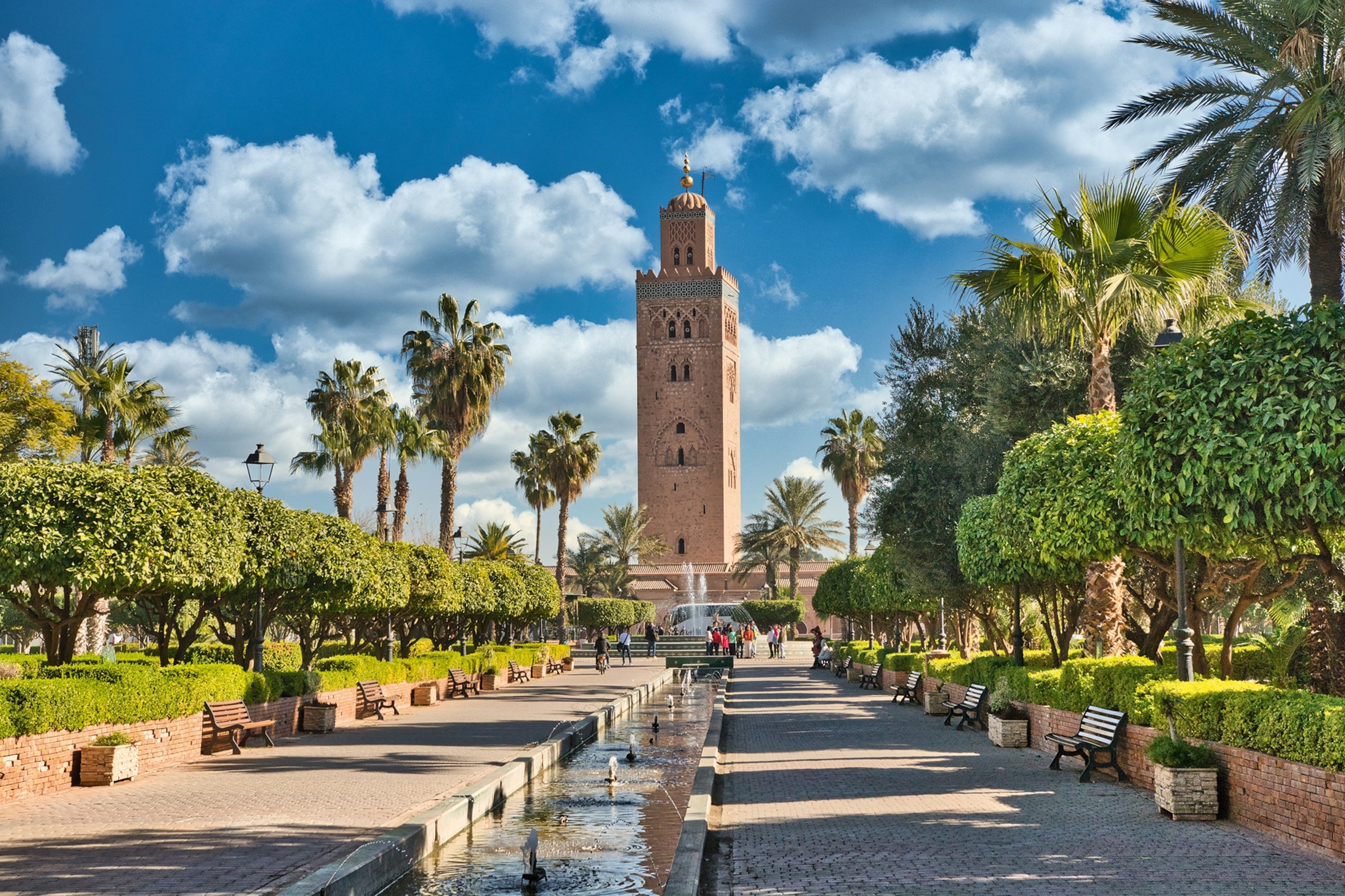 A palm-tree ringed plaza with a long fountain leading to a stone tower.