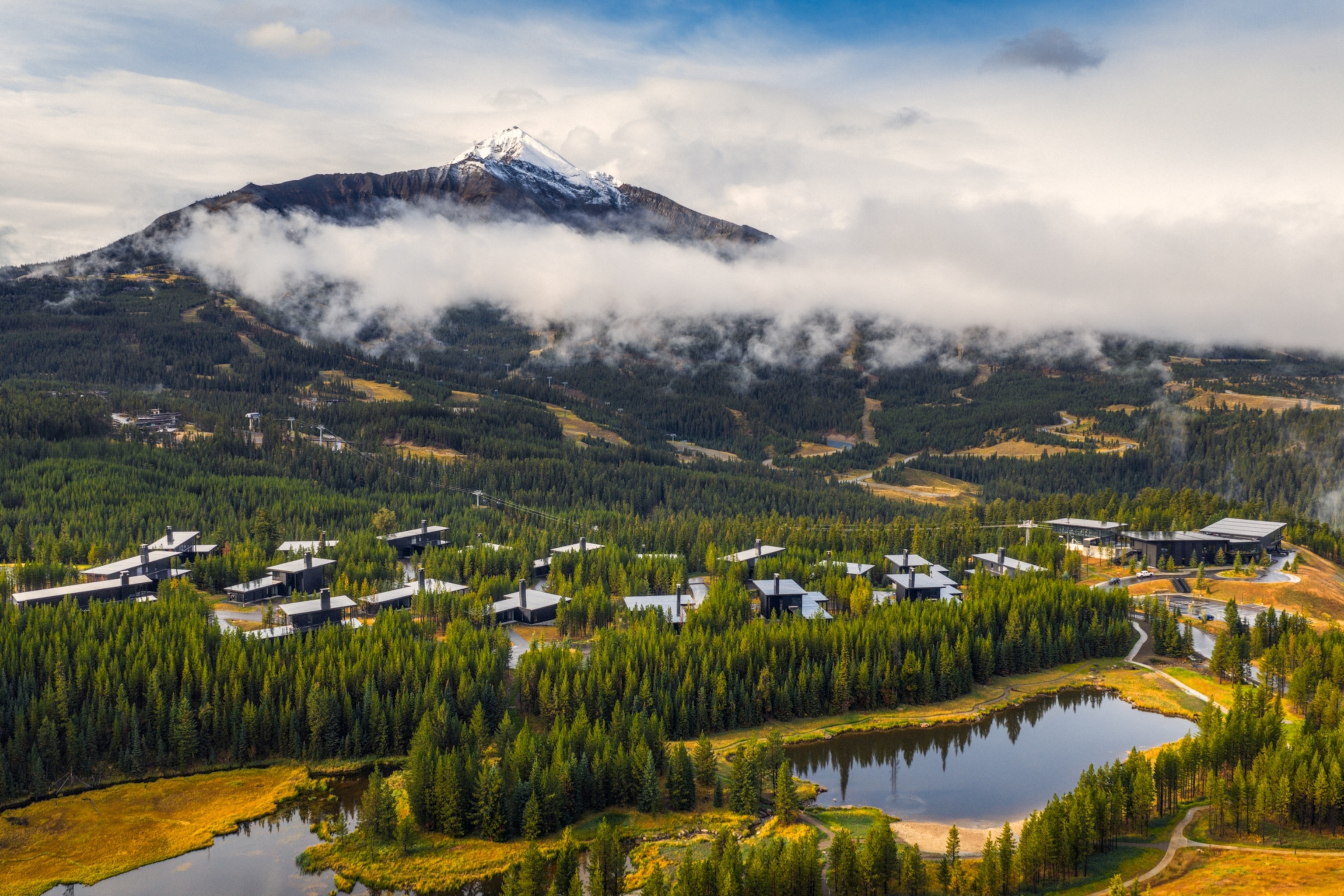 An aerial view of a mountain resort surrounded by pine forest, with a snow-capped peak rising above low clouds and small lakes in the foreground.