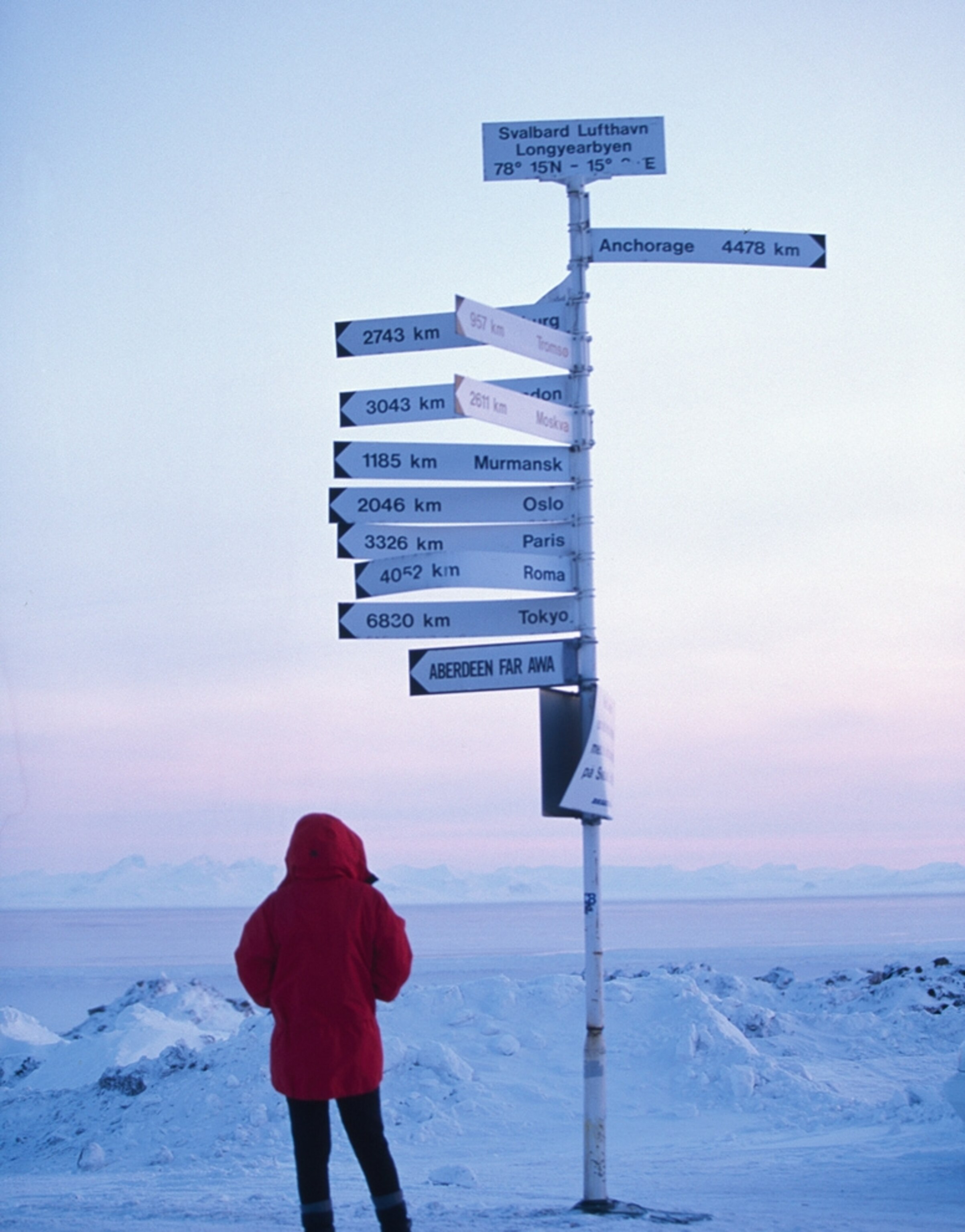 Northernmost airport picture: A person stands bundled up beside signs pointing south from Svalbard Airport in Longyear, Norway, for a gallery on the world's most extreme airports