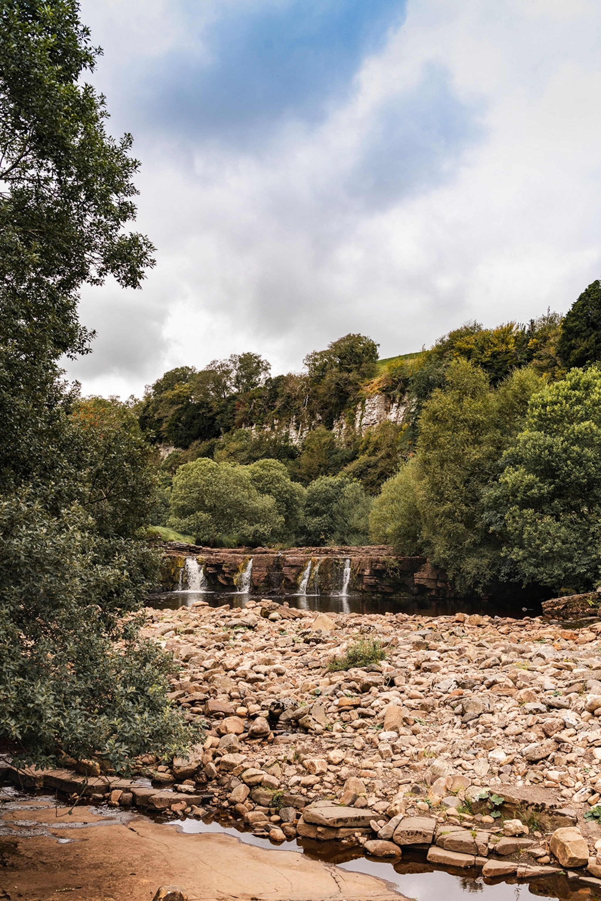 A shallow waterfall in a hill-side forest with water splashing onto gravel stones.