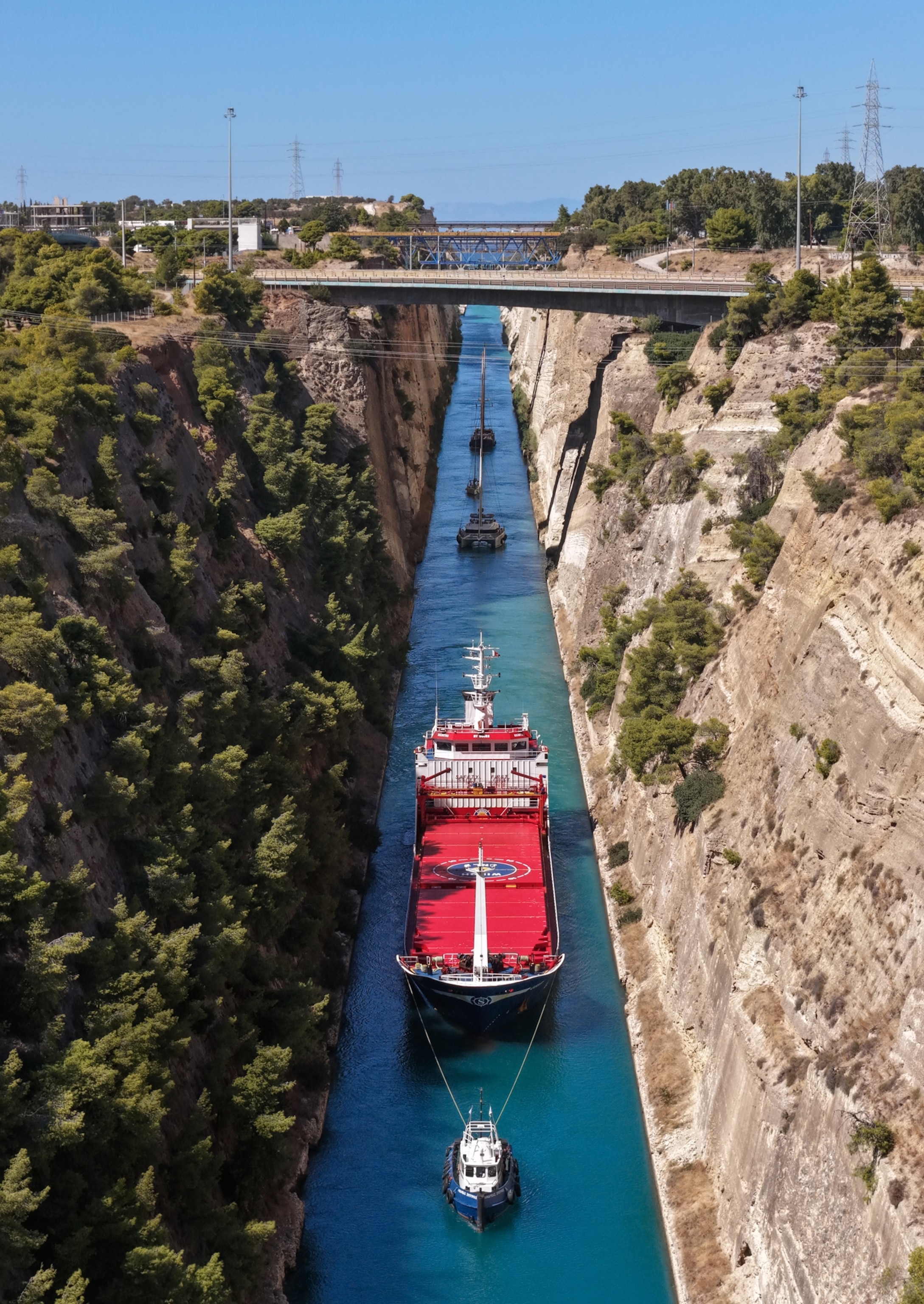 A large red cargo ship is guided by tugboat through the Corinth Canal