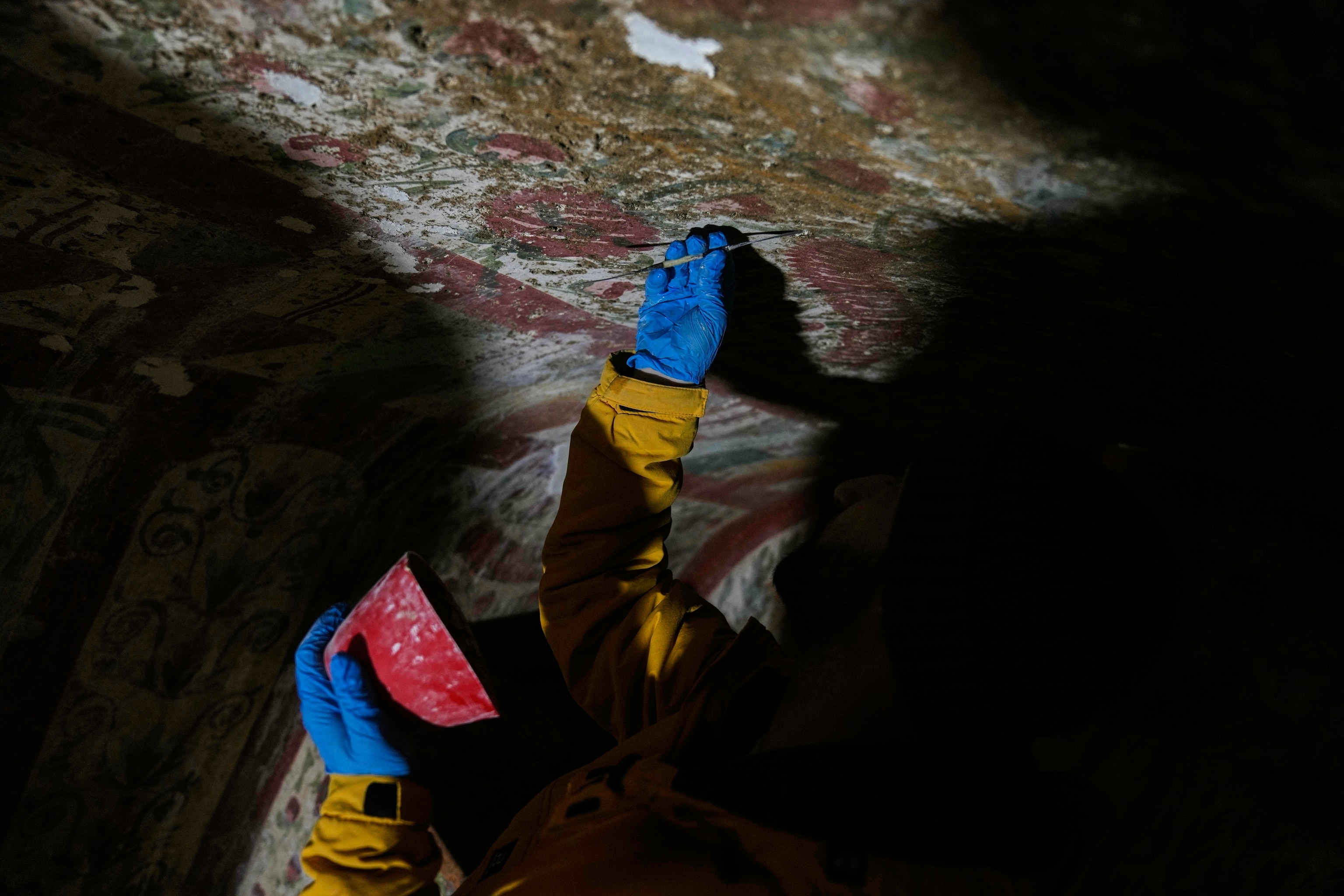 Gloved hands reach for the top of a tunnel a red bowl in the left hand and a small pick tool in the right hand, which is scraping the ceiling