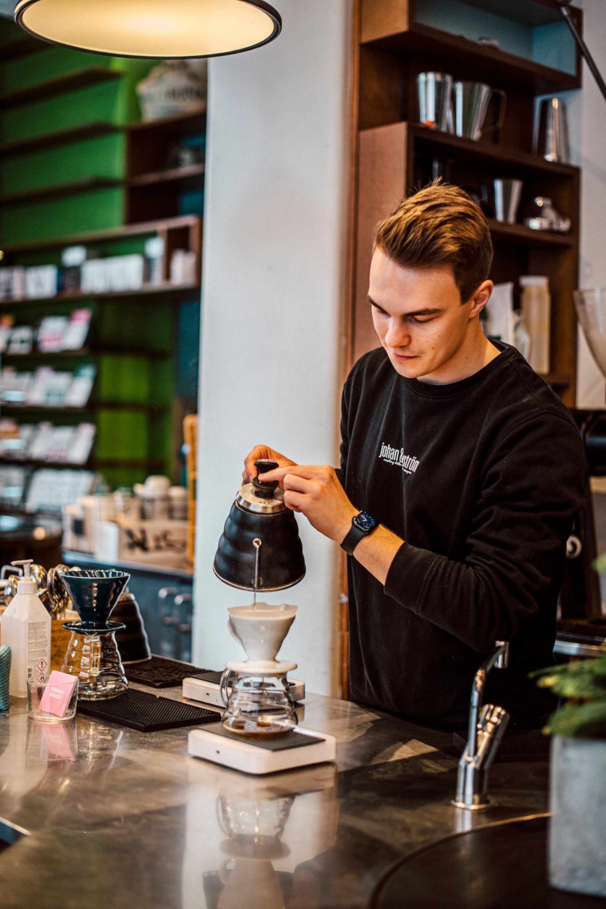 A barista in a cafe, pouring coffee into a filter that's resting on a cup on the countertop.