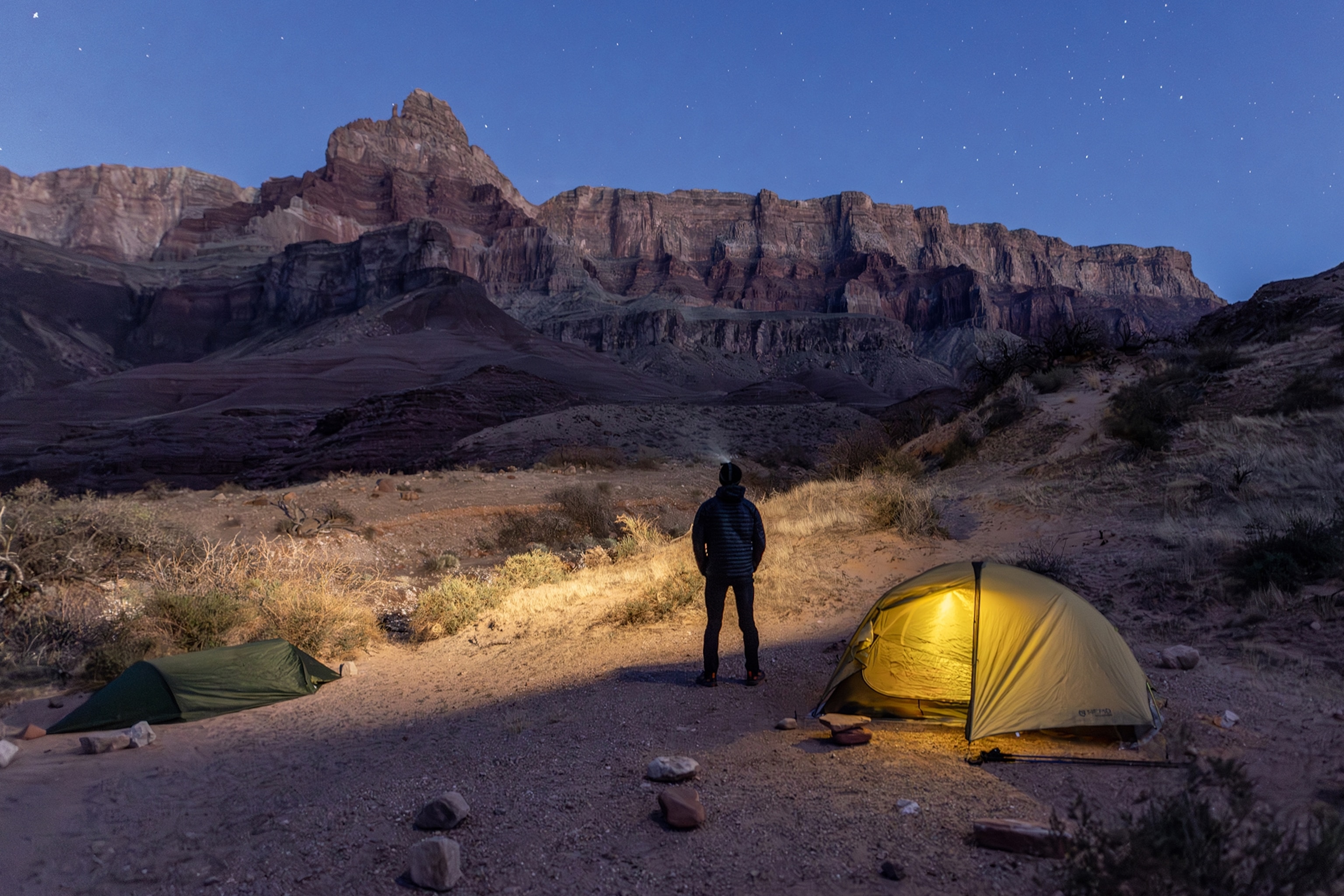 A man standing in front of his illuminated tend on the edge of a canyon at night.