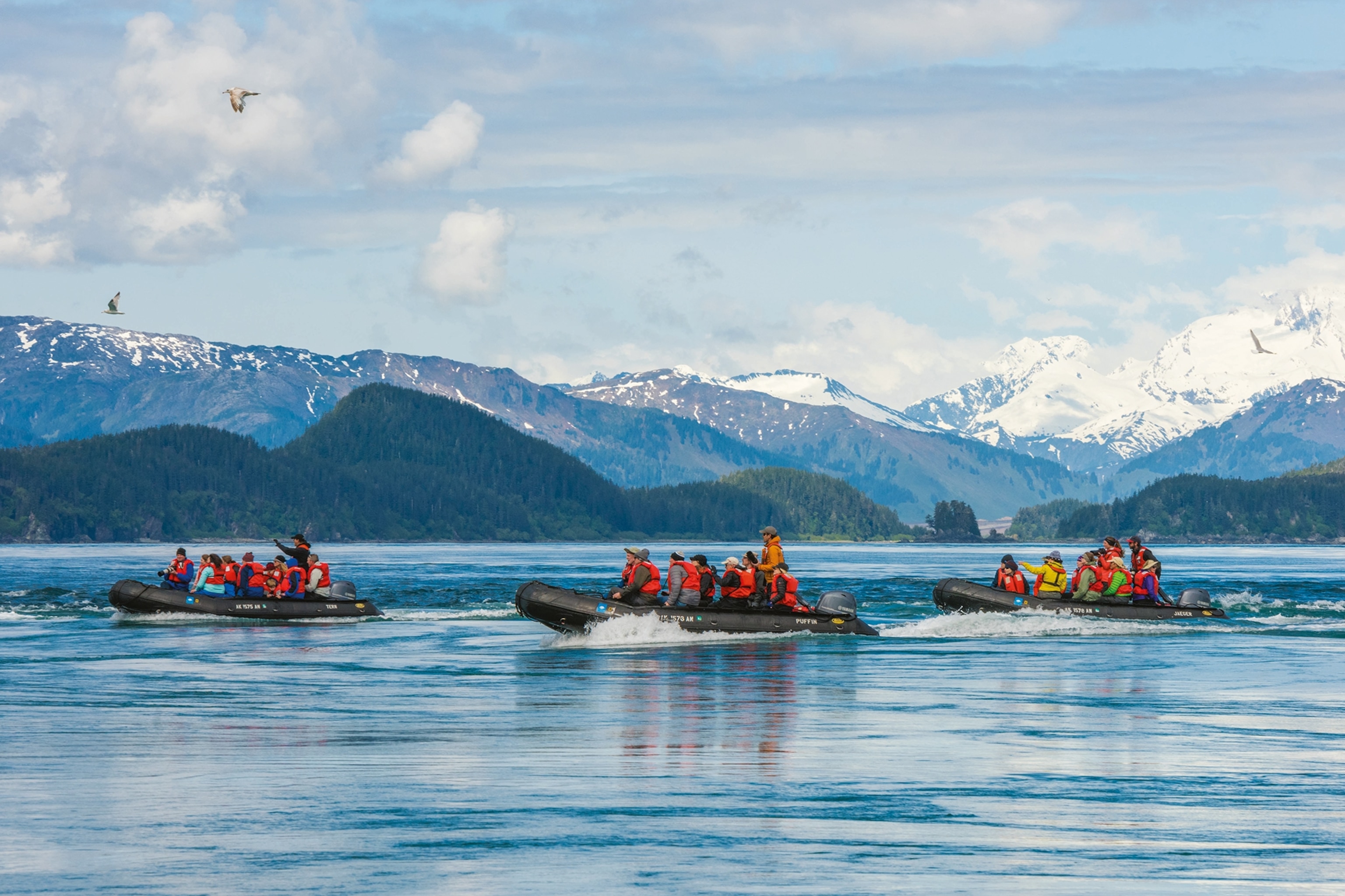 Three rubber boats with groups of visitors life jackets on a lake with snow-tipped mountains in the background.