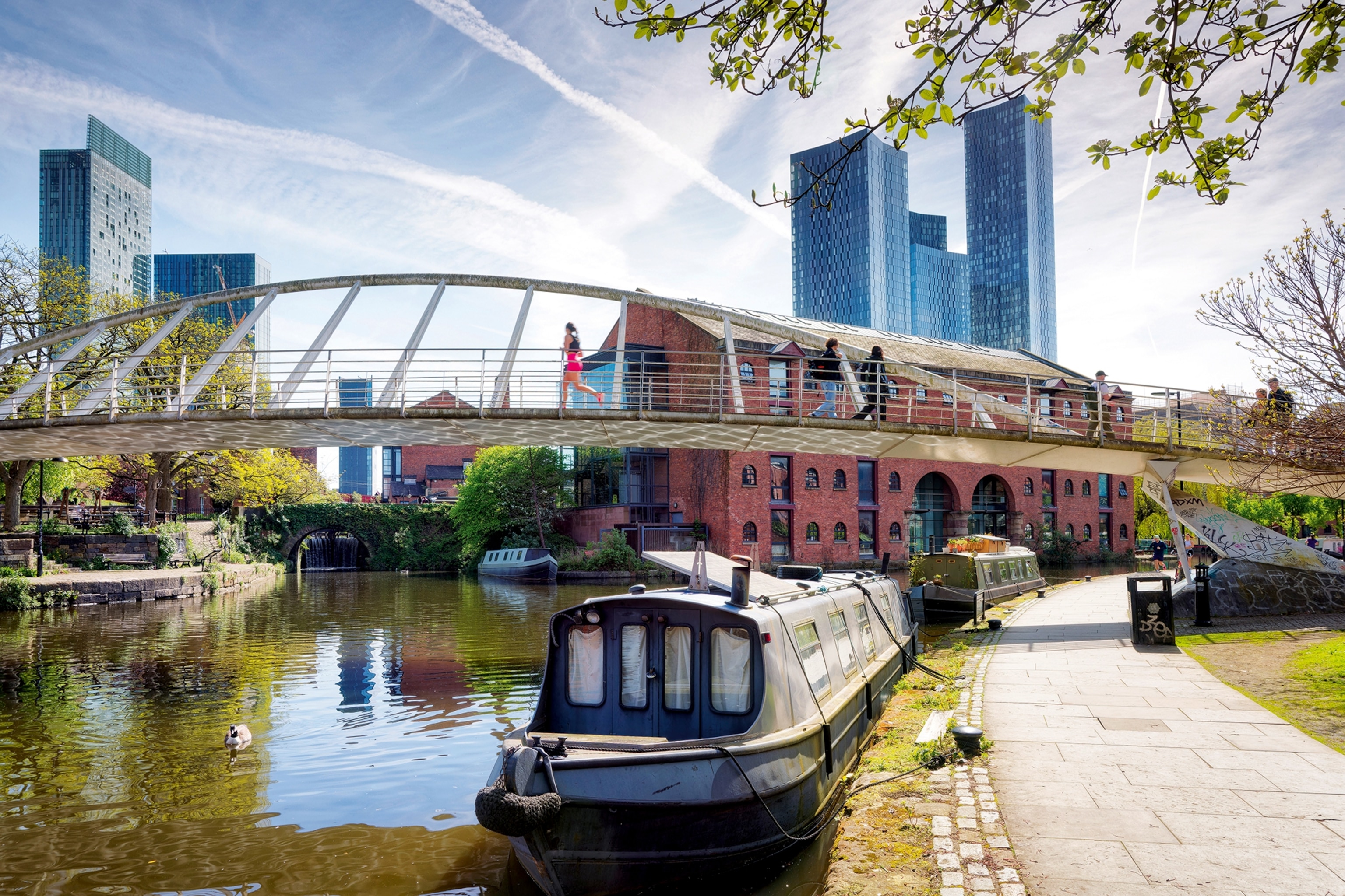 A canal in Manchester with an anchored boat and a bridge and skyscrapers in the background.