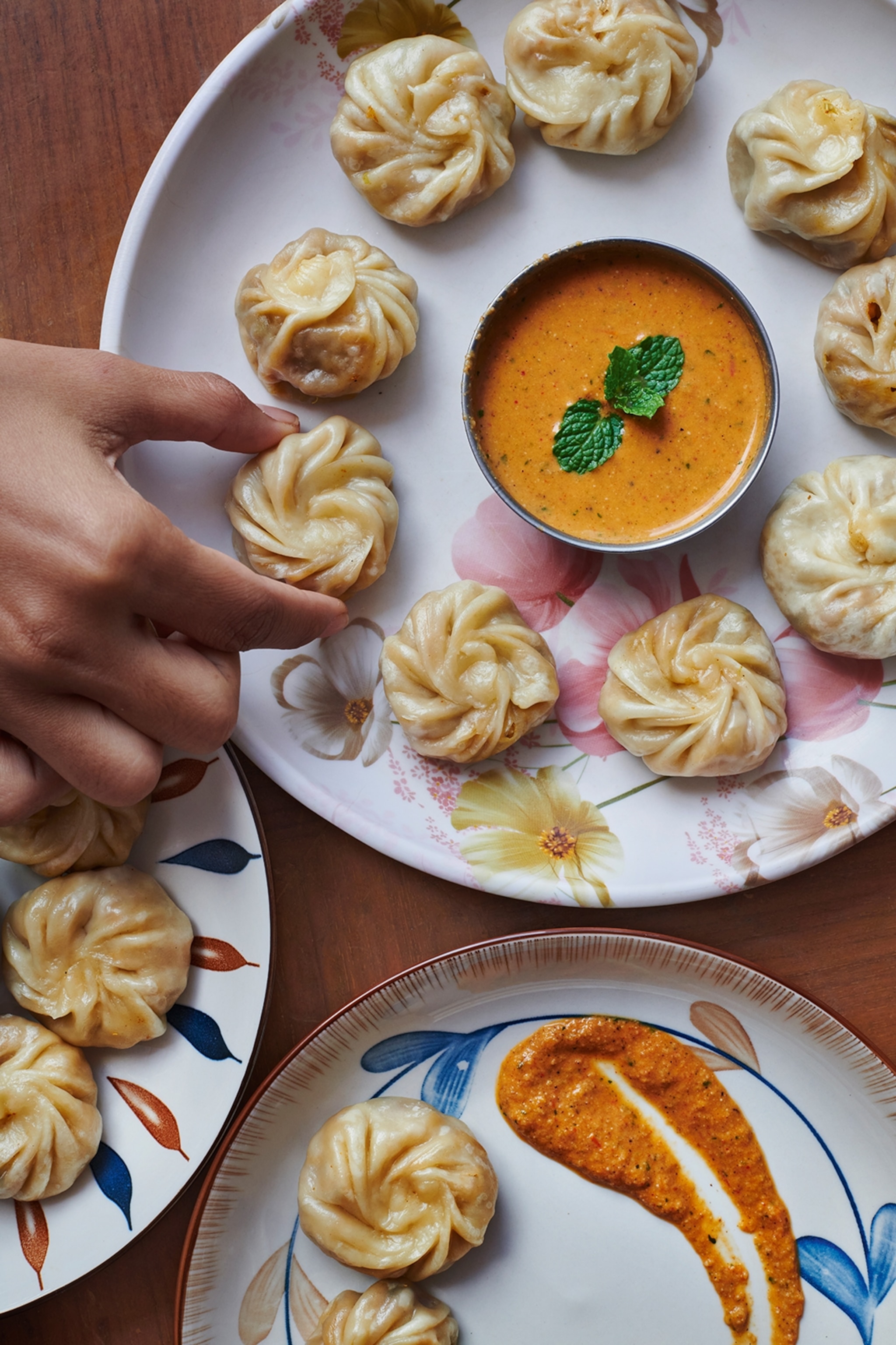 A close-up of a nine dumplings on a large plate with a spiced sauce in the middle and a hand picking up one.