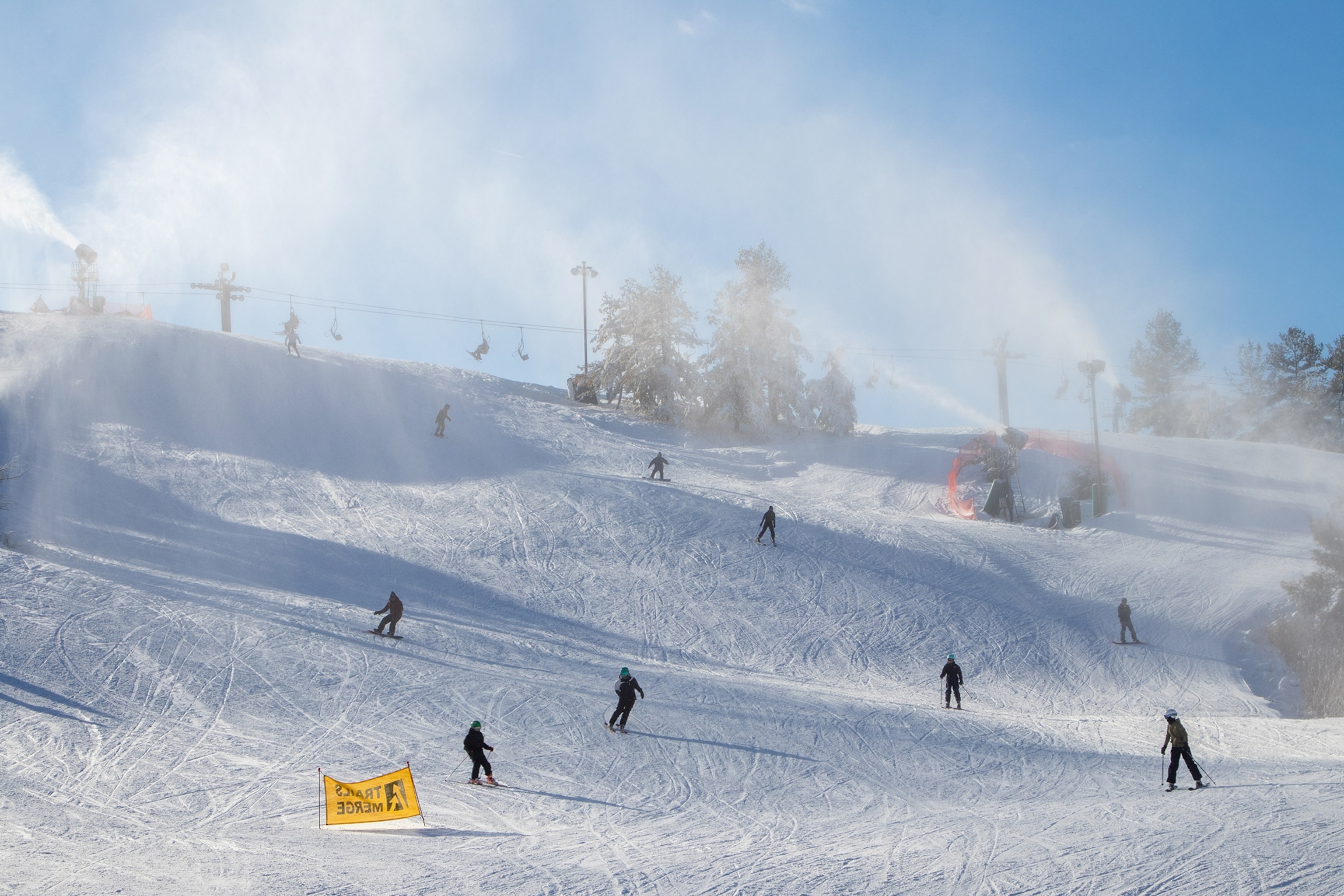 Snow cannons spray snow over a groomed skii run, several skiers and snowboarders on the slopes. A ski lift in the background.