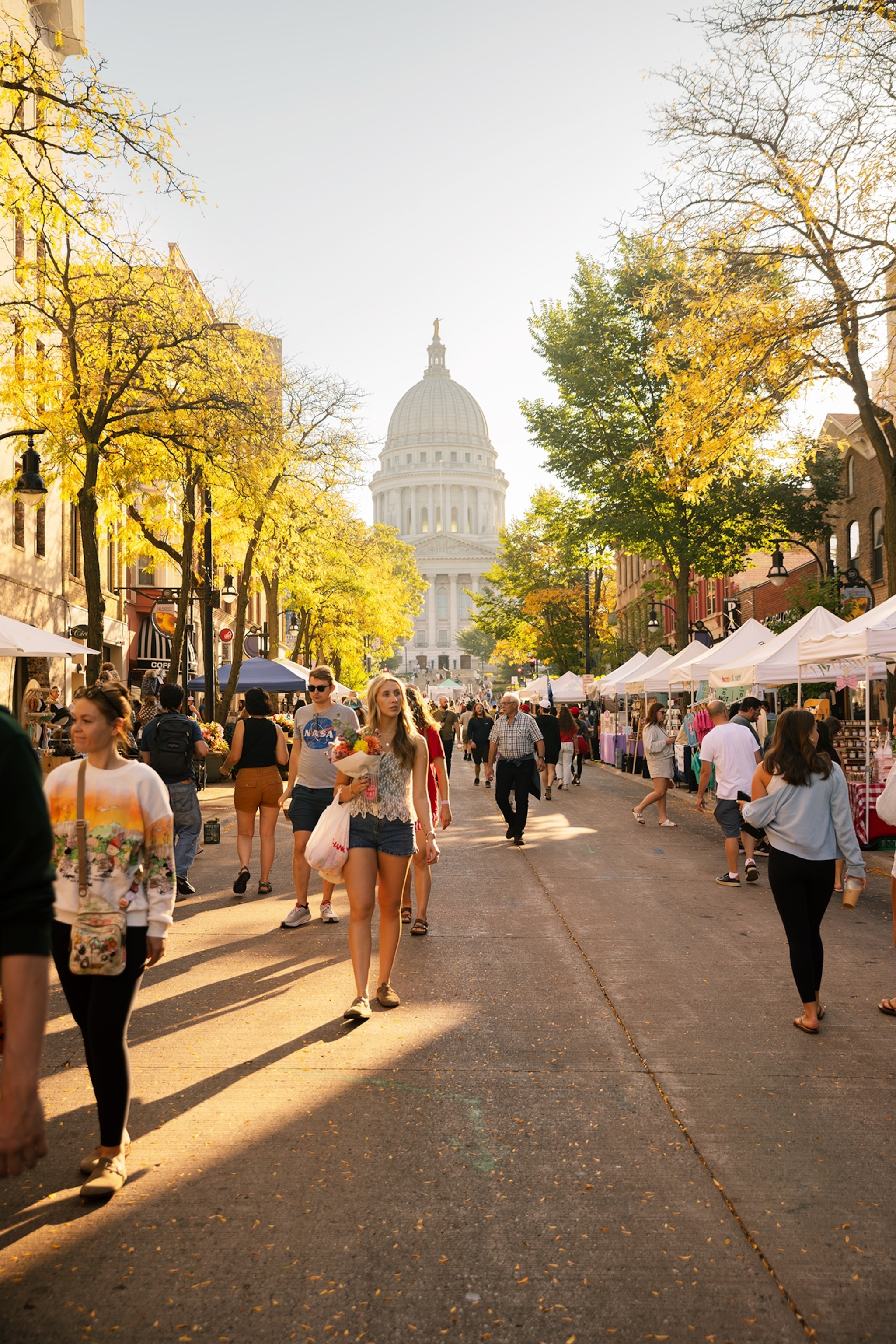 People walk through a busy outdoor market lined with white vendor tents, with a domed government building visible at the end of the street.