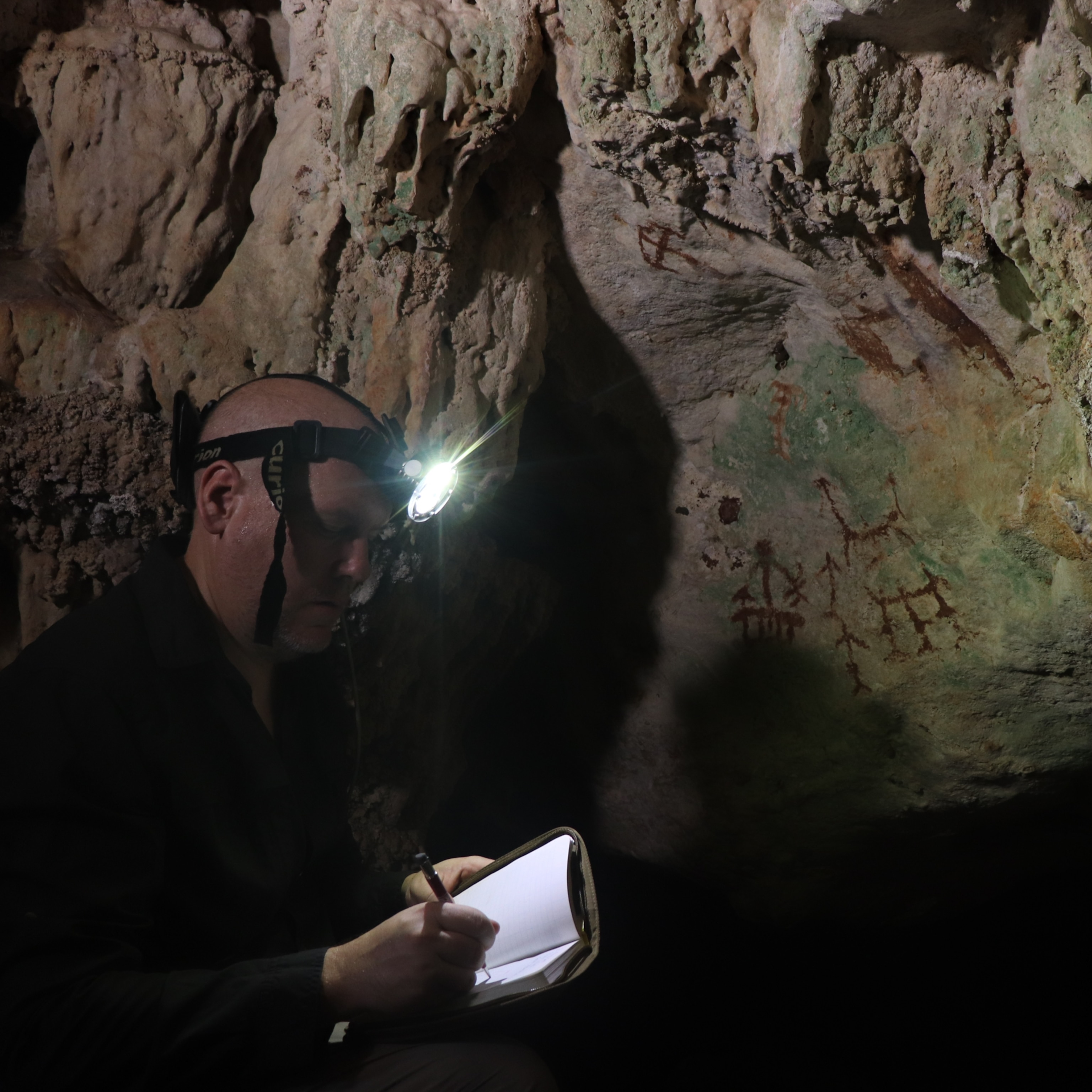 A man with a headlamp takes notes in a dimly lit cave. Ancient red cave paintings of animals and symbols cover the rocky walls.