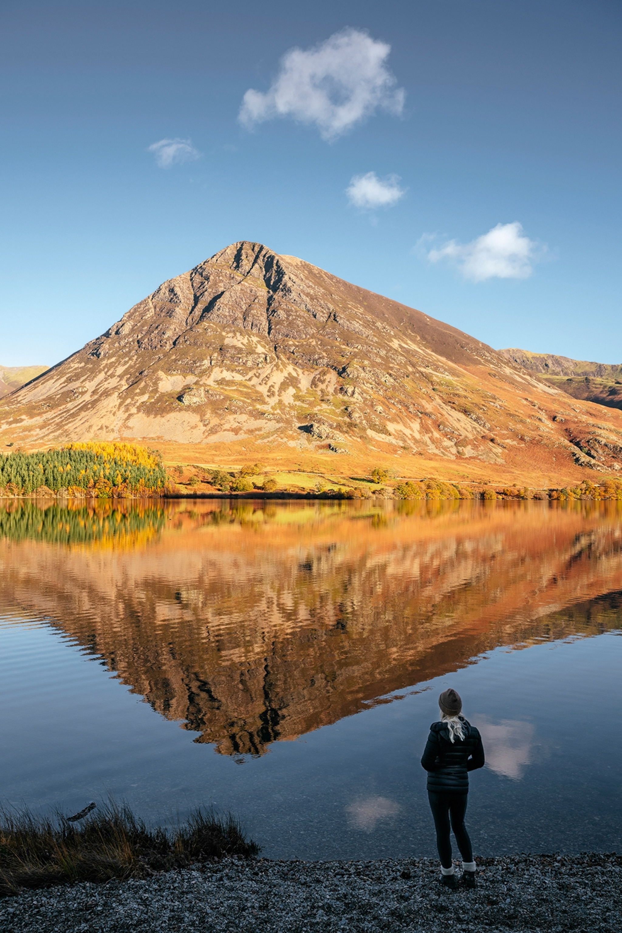 A hill reflected in a lake below with a hiker taking in the scene at its shore.